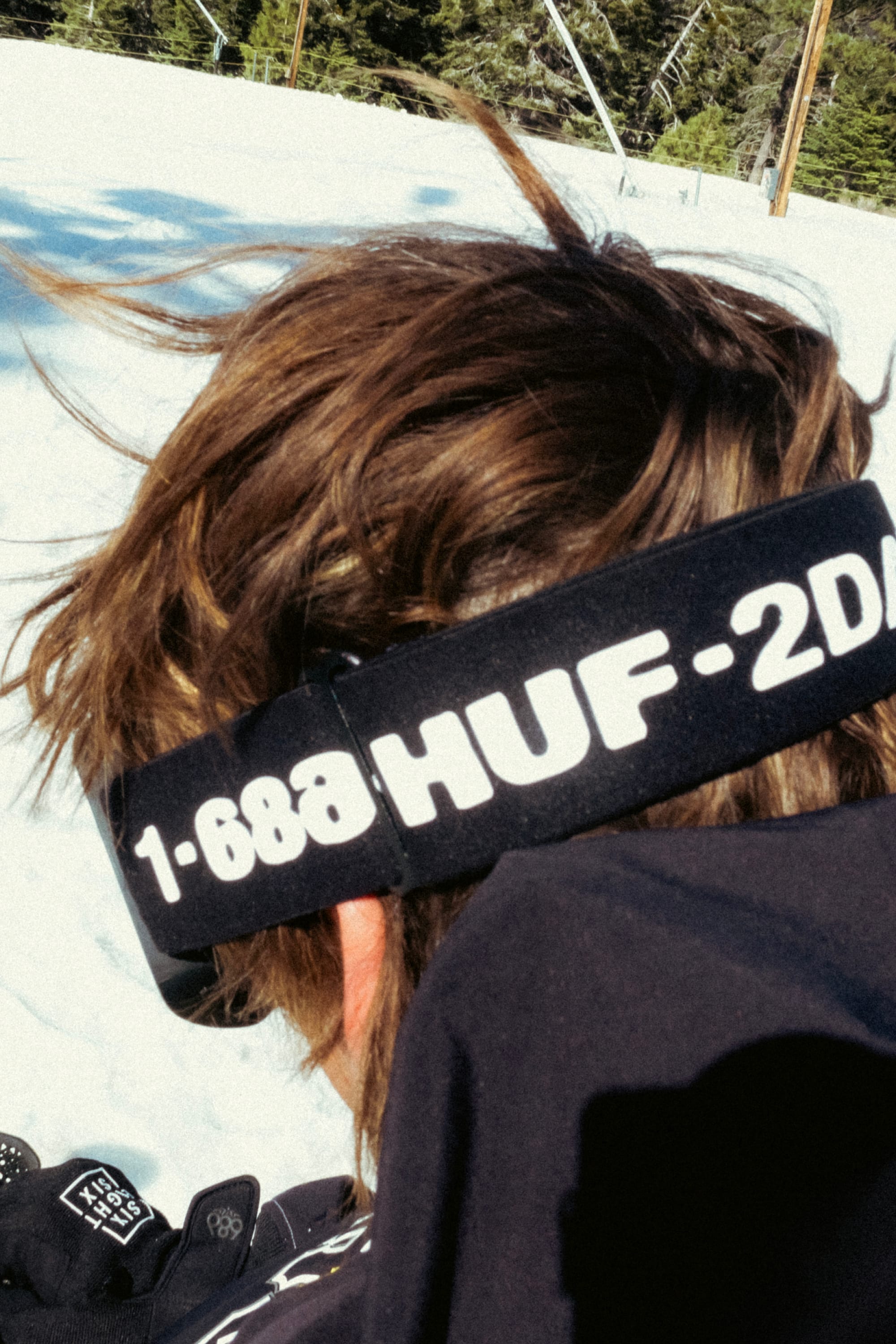 A person with brown hair wears the 686 SMARTY Goggle with minimalist design and magnetic lenses, branded 686 on the strap, standing on snow with trees and a blue sky—ideal for all conditions.