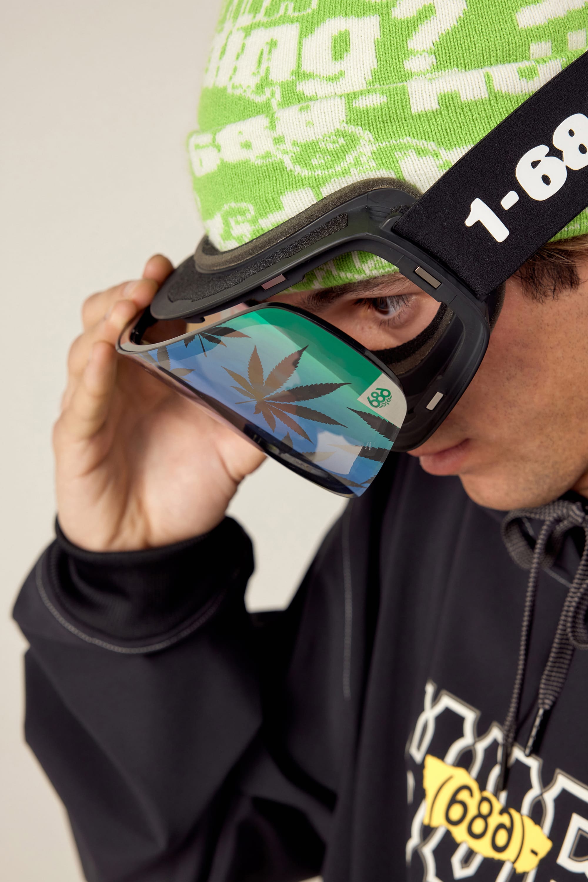 A person in a green beanie and black hoodie lifts the 686 SMARTY Goggle, reflecting a marijuana leaf on the lens. The headband shows 1-68 and the beanie displays white graphics—perfect for all conditions.
