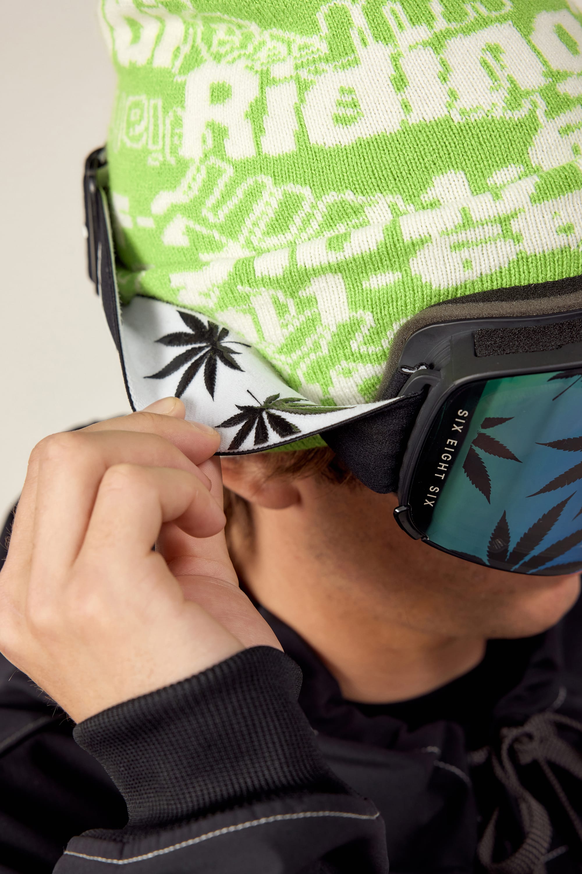 Wearing the 686 SMARTY Goggle with magnetic blue-tinted lenses and a green patterned beanie, a person adjusts a white neck gaiter featuring black marijuana leaves. Only part of their face is seen, spotlighting 686’s all-conditions gear.