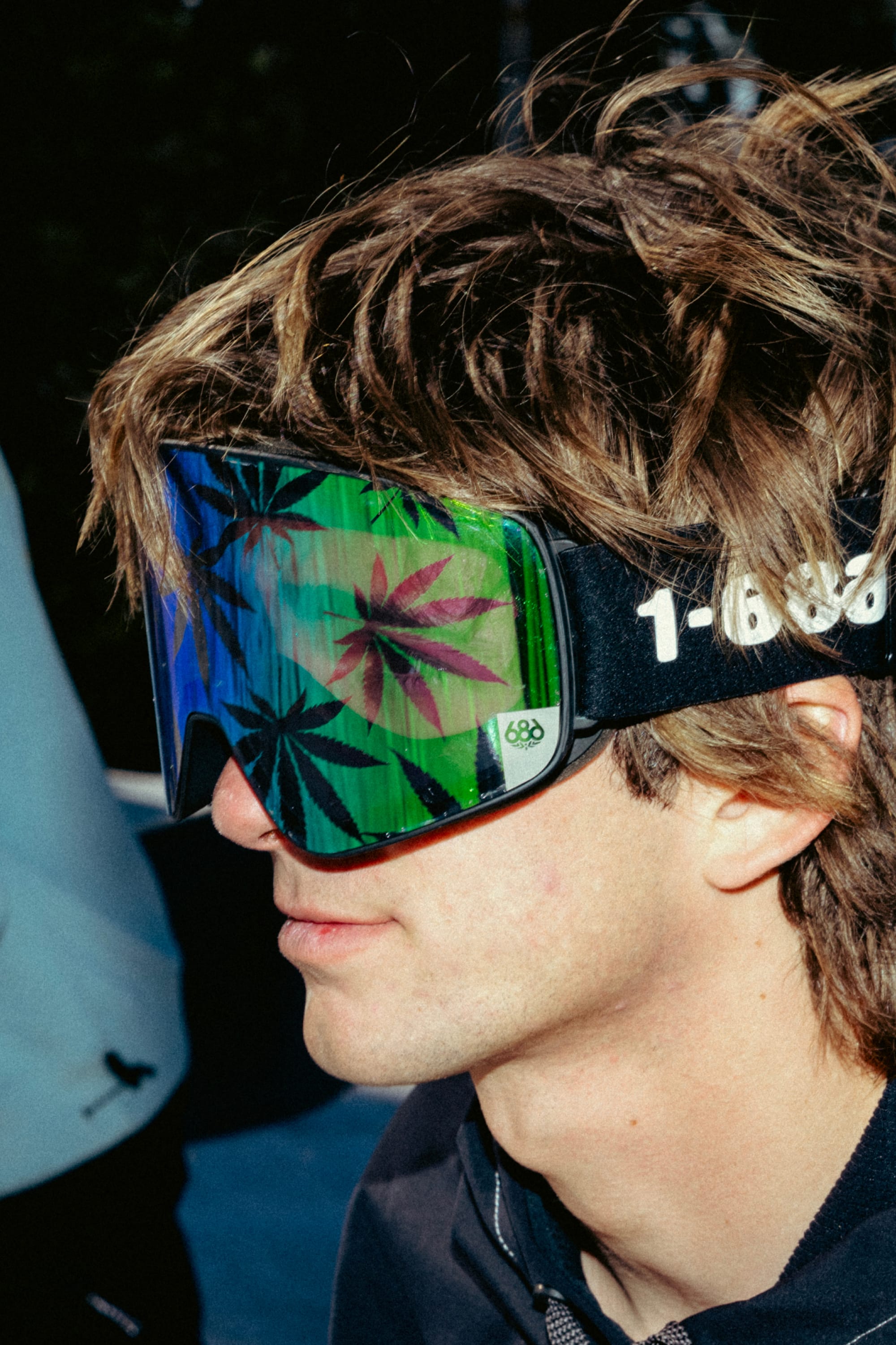 A person with brown, tousled hair wears 686 SMARTY Goggle by 686—large minimalist goggles with magnetic lenses reflecting green marijuana leaves—paired with casual outdoor clothing for all conditions.