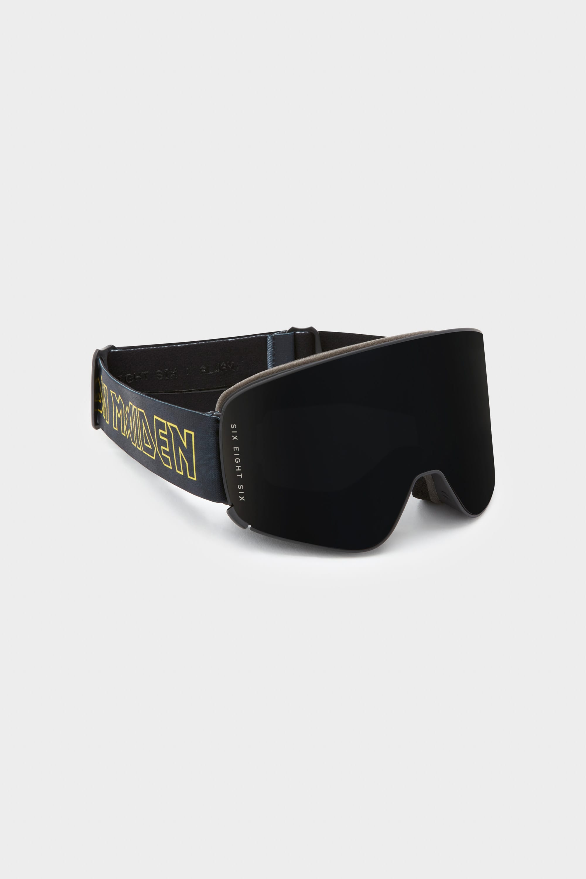 The 686 SMARTY Goggle by 686 features a sleek black minimalist design, curved magnetic lenses for easy swaps in any condition, an adjustable strap with bold yellow IRON MAIDEN text and logo, all showcased against a white background.