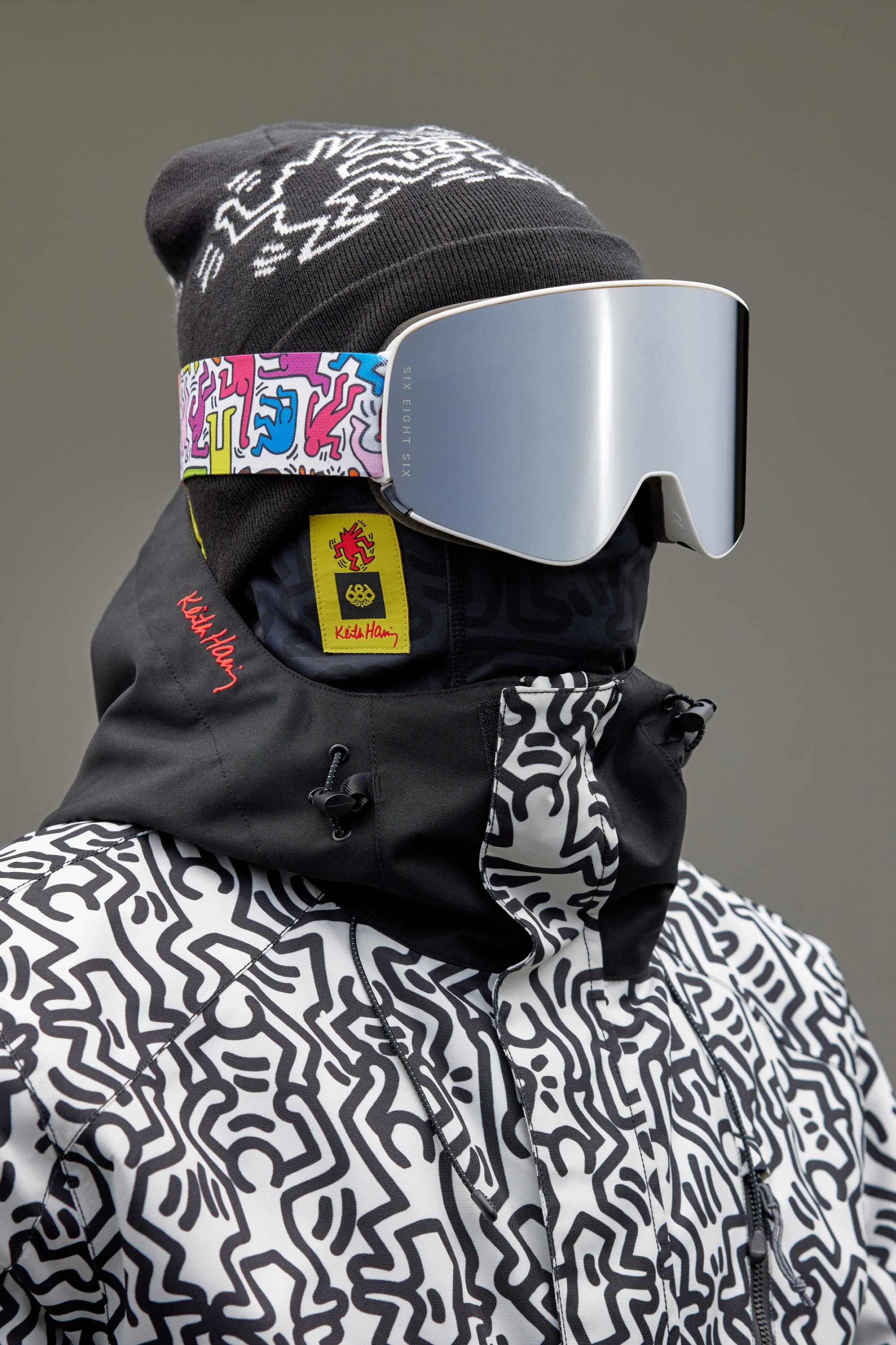 Wearing 686 SMARTY Goggle ski goggles with magnetic lenses and a minimalist design, along with a black beanie and Keith Haring-style patterned jacket—both featuring red Keith Haring tags. Gray background. Brand: 686.