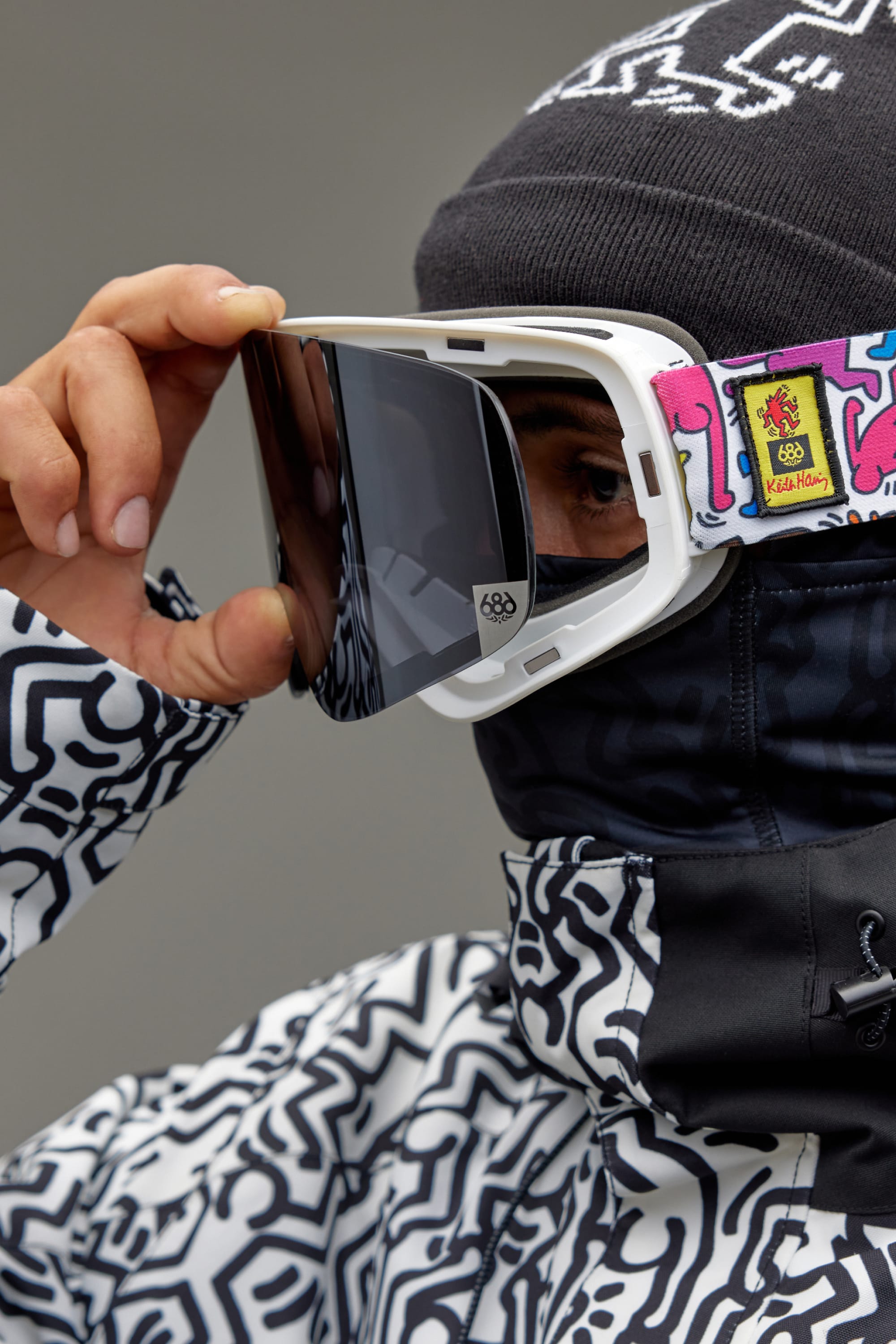 Wearing patterned ski gear and a black beanie, a person adjusts their 686 SMARTY Goggle by 686, featuring minimalist magnetic lenses and a strap with colorful Keith Haring artwork. The plain background suggests theyre ready for any condition.