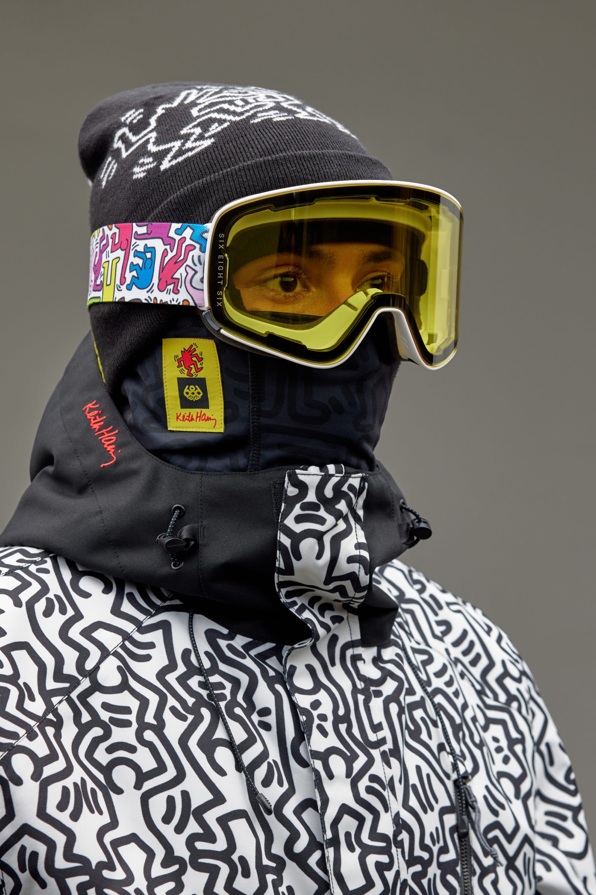 A person wears a black-and-white patterned jacket, matching face cover, dark beanie with colorful designs, and the 686 SMARTY Goggle by 686—yellow minimalist goggle with magnetic lenses and a boldly designed strap—all set against a plain gray background.