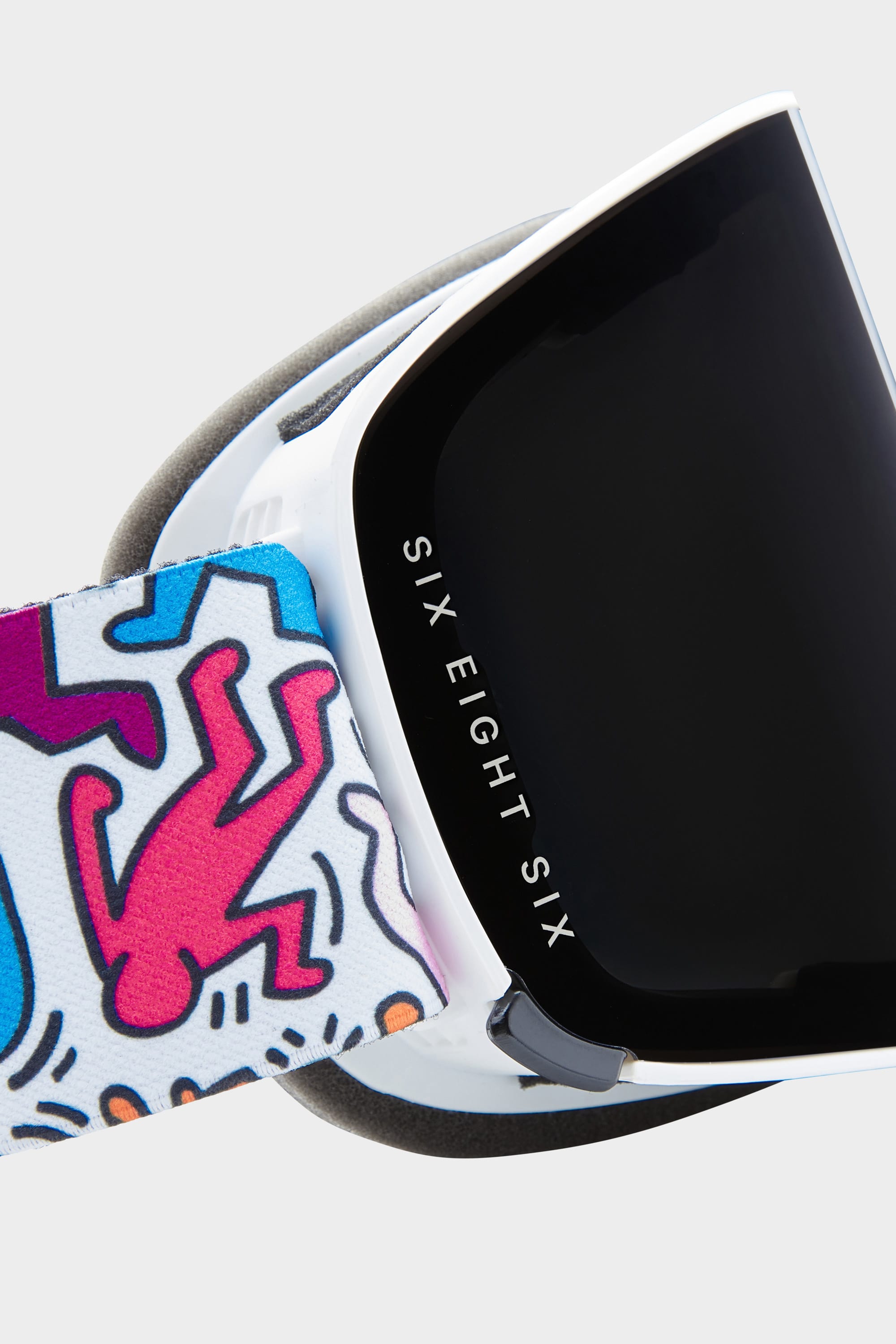 Close-up of the 686 SMARTY Goggle: minimalist design with a black lens, white frame, and a colorful strap featuring abstract pink and blue figures. 686 branding on the side; magnetic lenses provide reliable performance in all conditions.