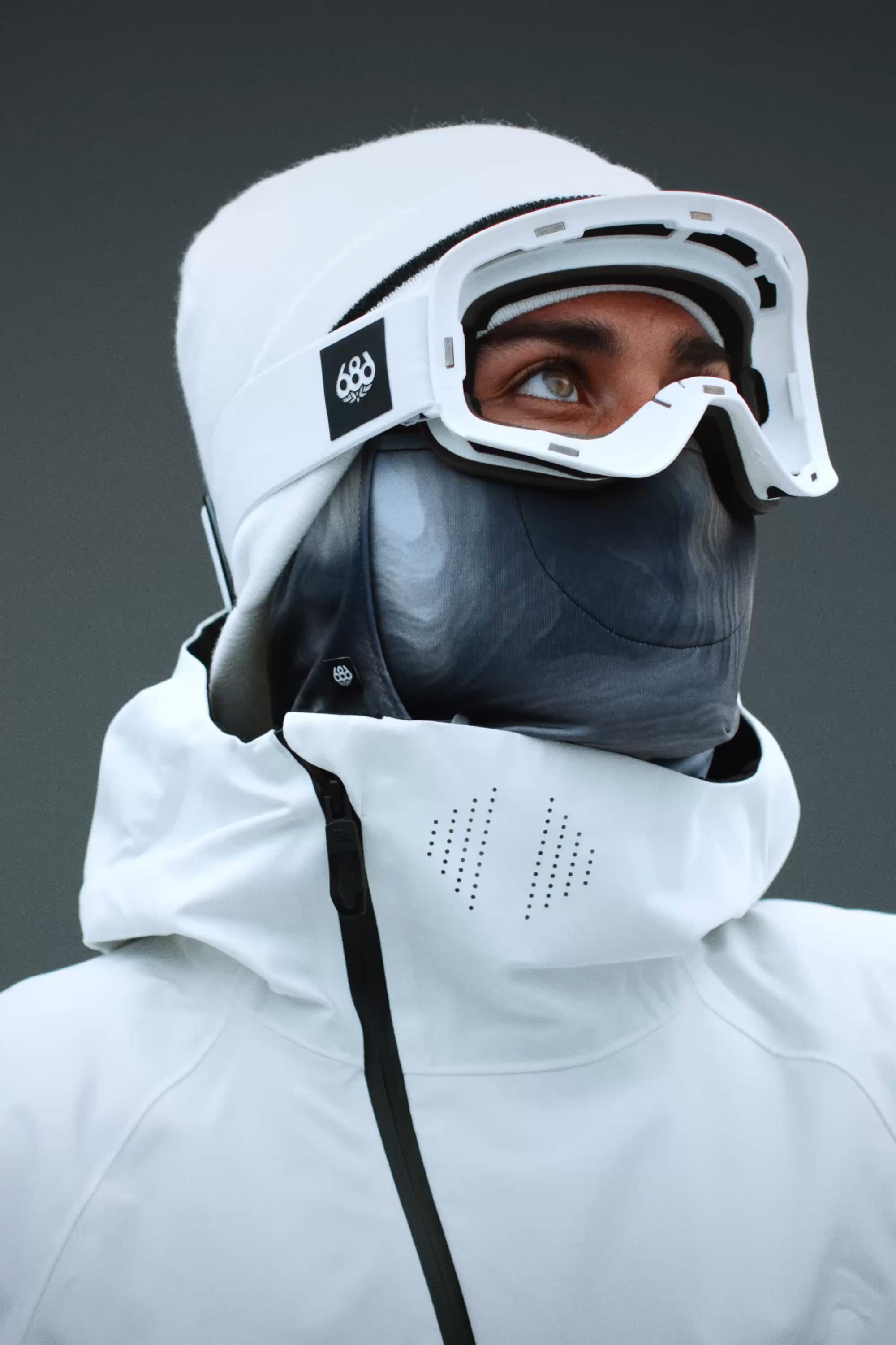 A person wearing a white jacket, white beanie, and the 686 SMARTY® Magnetic Goggle by 686 gazes upward. Their face is mostly concealed by a dark mask, set against a plain grey background.