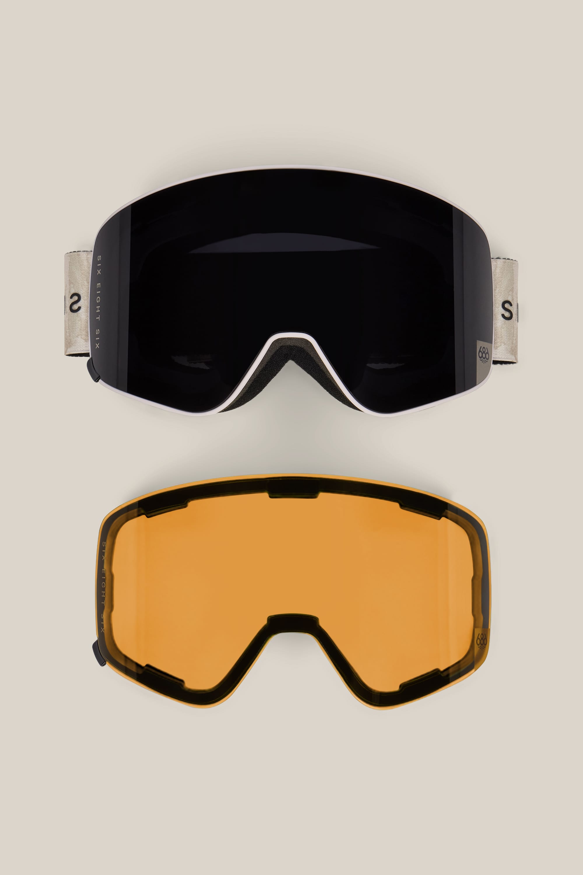 The 686 SMARTY® Magnetic Goggle by 686, featuring a black lens and a beige strap, is displayed above a separate yellow replacement lens on a plain beige background.