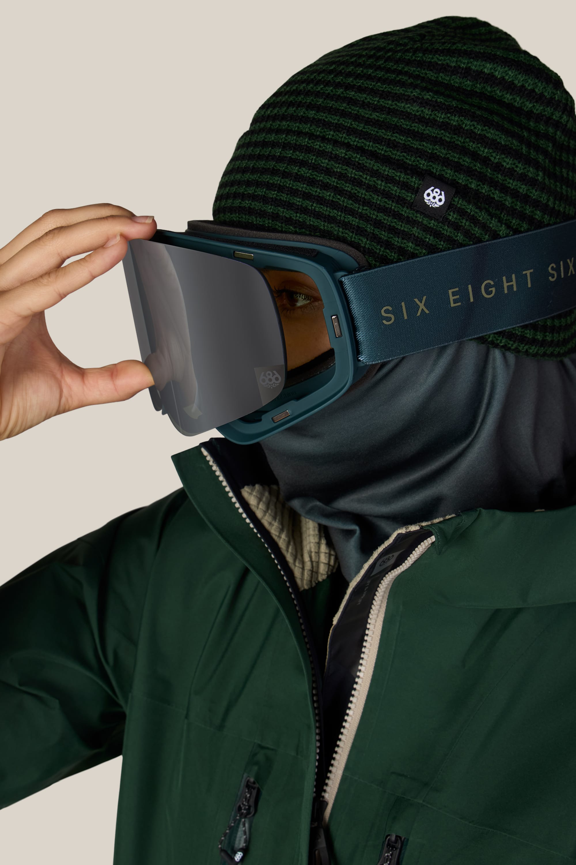 A person wearing a green jacket, striped beanie, face mask, and reflective ski goggles labeled “686” on the strap is adjusting their 686 SMARTY® Magnetic Goggle with one hand.