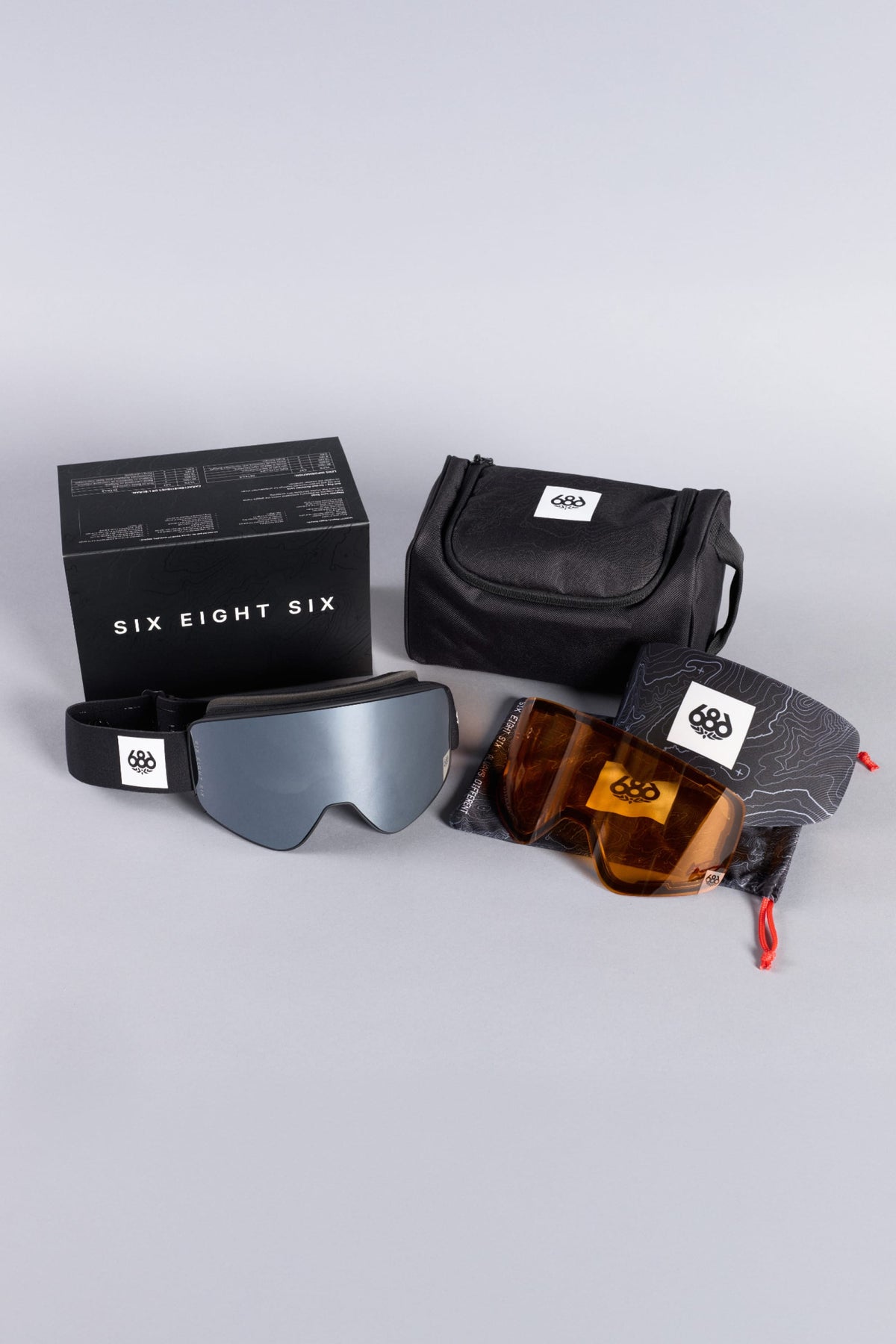 The 686 SMARTY® Magnetic Goggles by 686, featuring interchangeable lenses, come with a black carrying case, a patterned cloth pouch, and are presented in a black box labeled “Six Eight Six” on a light surface.