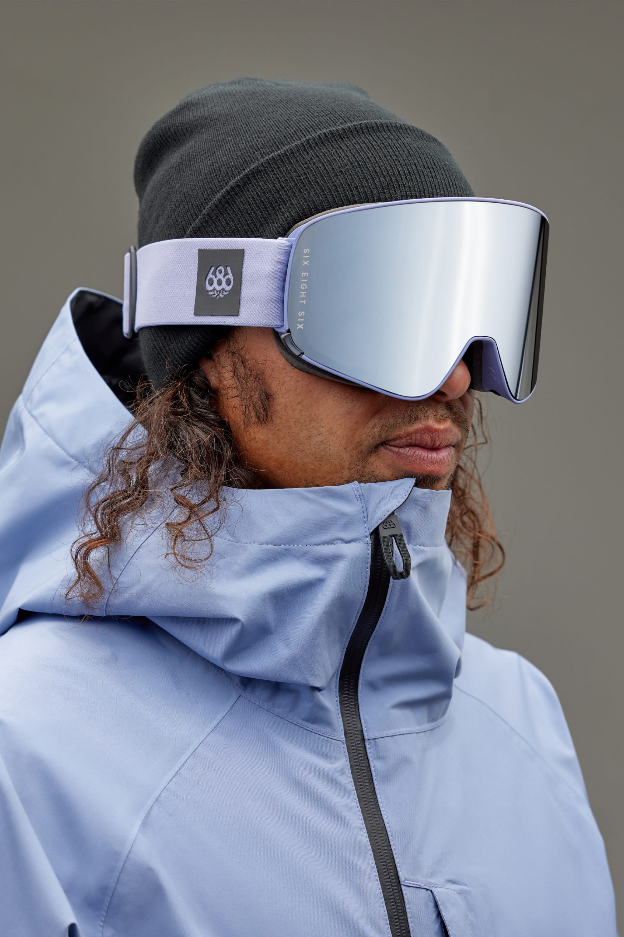 A person wearing the 686 SMARTY Goggle, a black beanie, and a light blue winter jacket stands against a plain gray background. Their curly hair peeks out below the beanie, ready for all conditions.