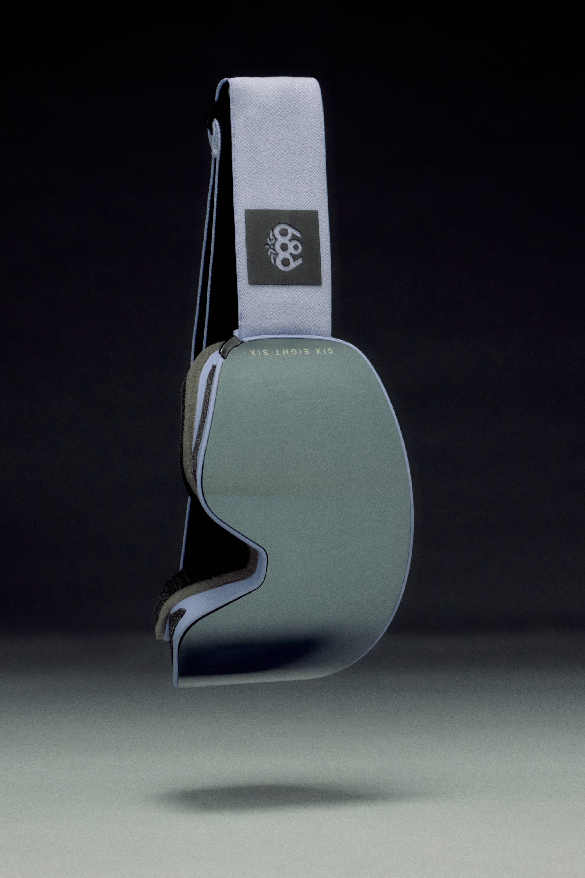 The 686 SMARTY Goggle by 686 features a minimalist reflective lens and white strap with a black patch sporting the white four-knuckle logo, all designed for all conditions and displayed against a dark gradient background.
