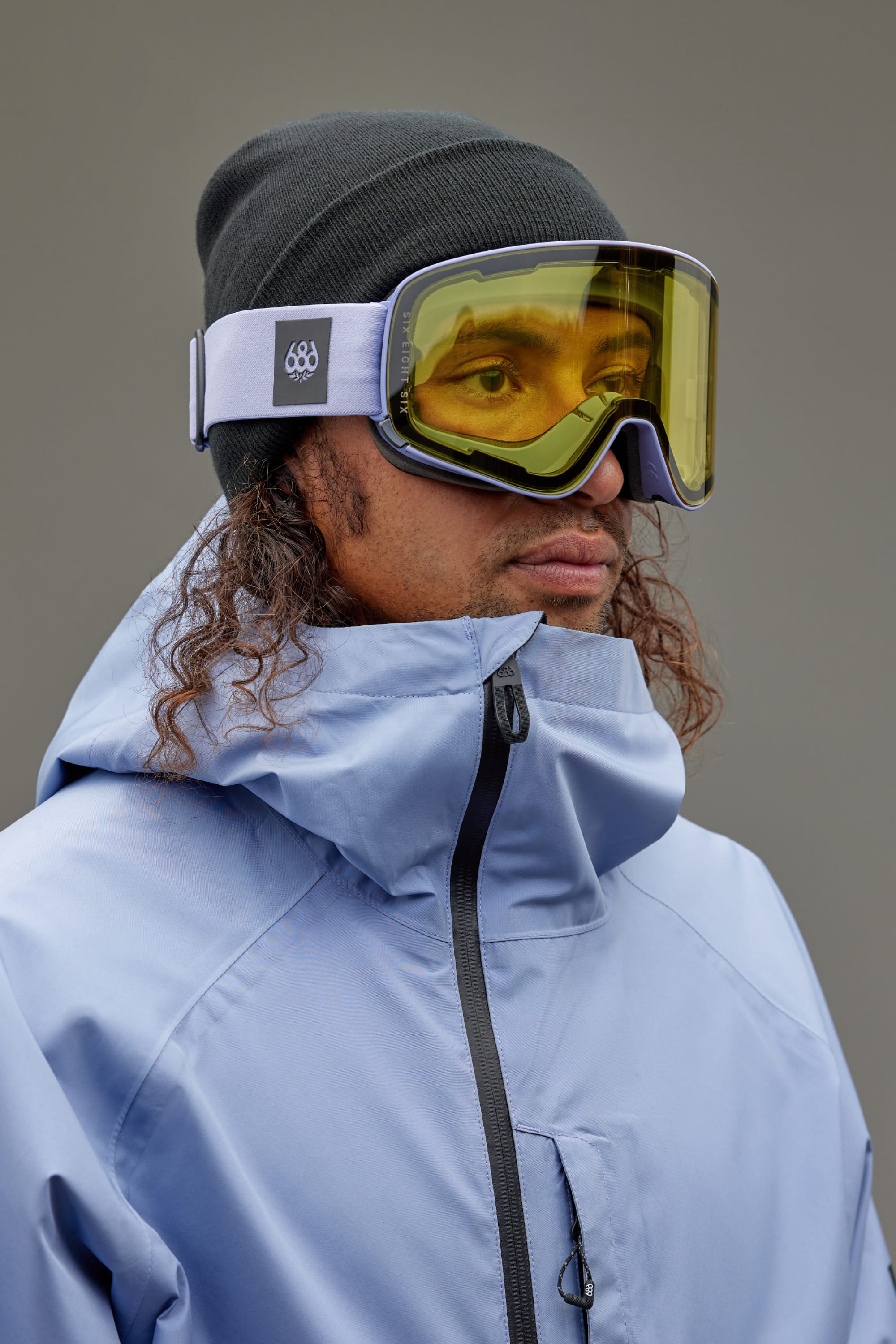 Wearing a black beanie and a light blue high-collar jacket, a person stands against a gray background sporting the 686 SMARTY Goggle by 686, featuring minimalist design and magnetic lenses—ready for all conditions.