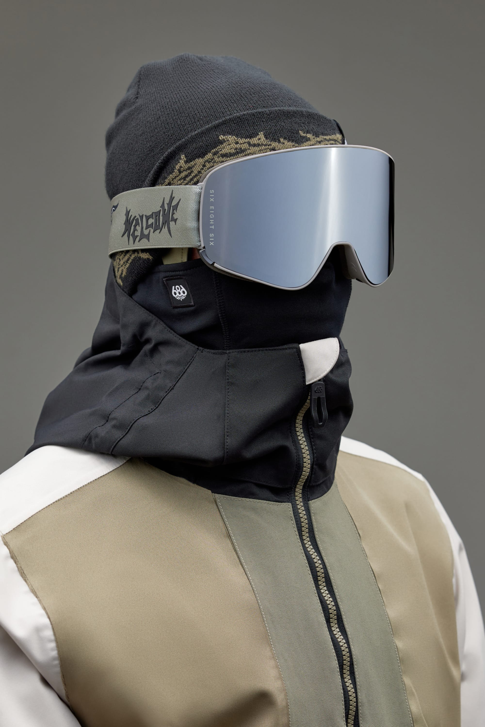 A person wears a black beanie, 686 SMARTY Goggle with magnetic lenses, a black face covering, and a beige, black, and white color-blocked jacket from 686, standing against a gray background—geared up for any condition.