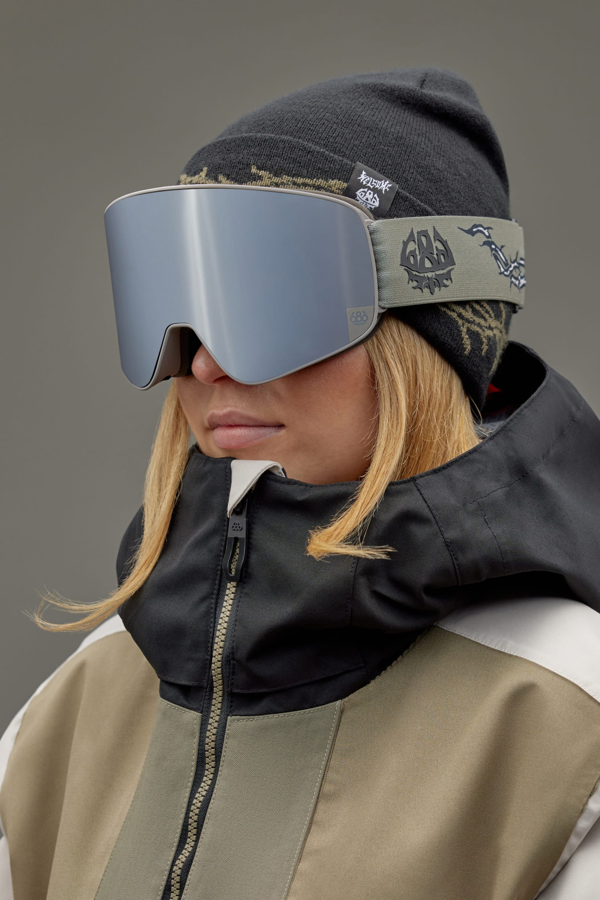 A person with long blond hair under a black beanie wears the 686 SMARTY Goggle by 686, featuring large magnetic lenses, plus a beige and black winter jacket, standing against a plain gray background.