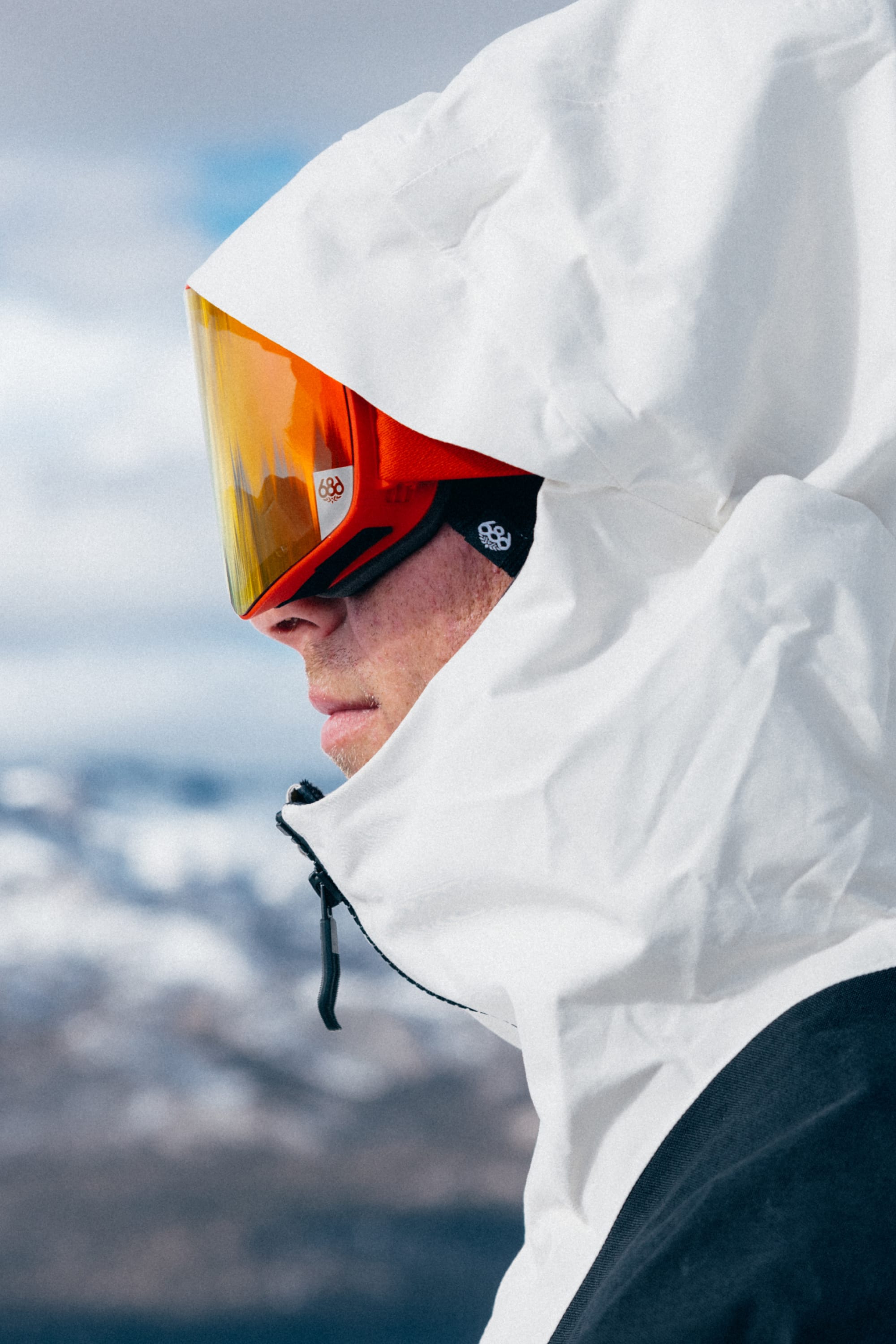 A person in a white winter jacket and 686 SMARTY® Magnetic Goggles by 686 stands outdoors, with snowy mountains visible in the blurred background.