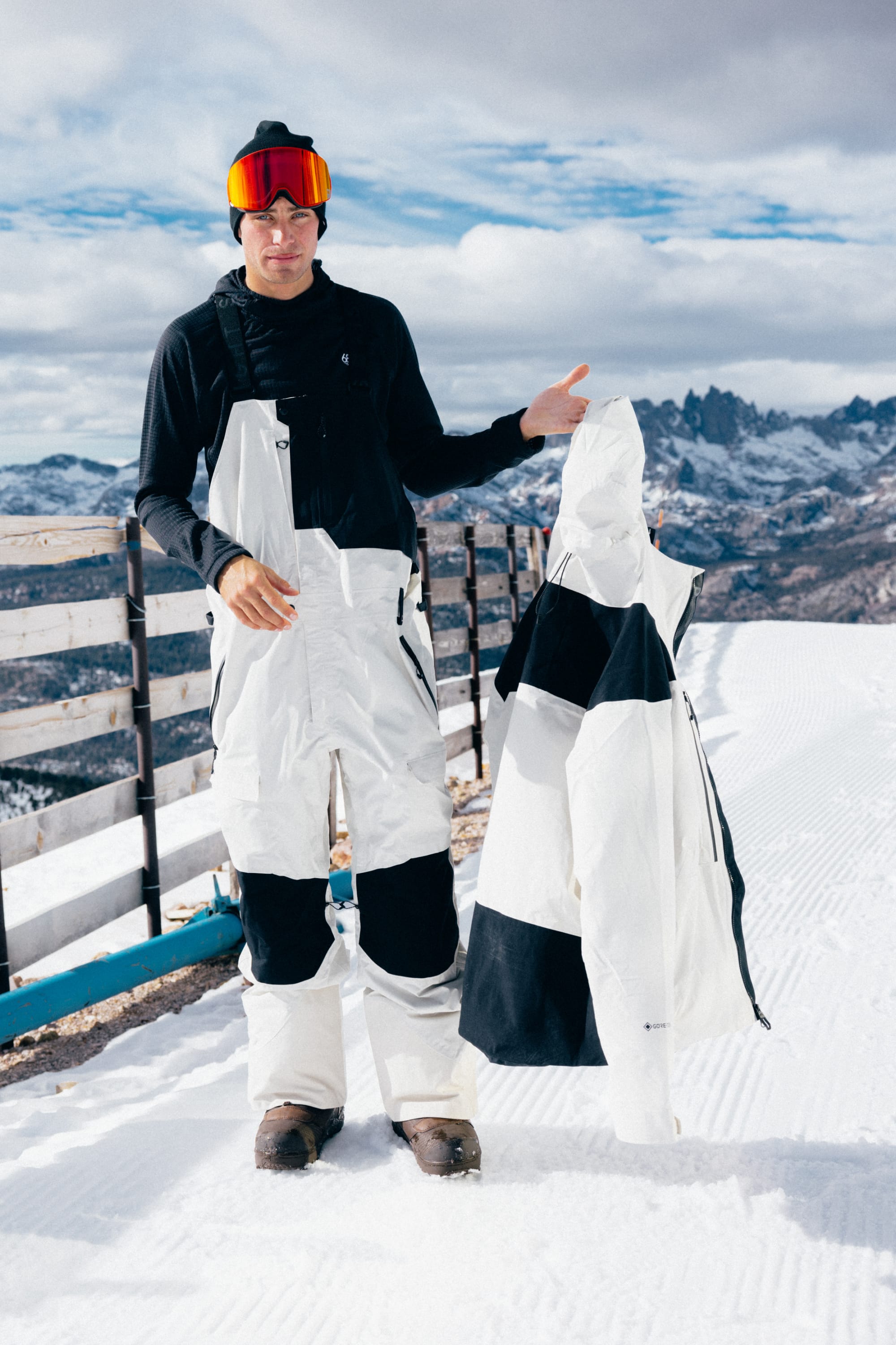 A skier dressed in white pants and a black shirt stands on a snowy mountain, wearing the 686 SMARTY® Magnetic Goggle by 686 and holding a white and black jacket, with mountains visible in the background.