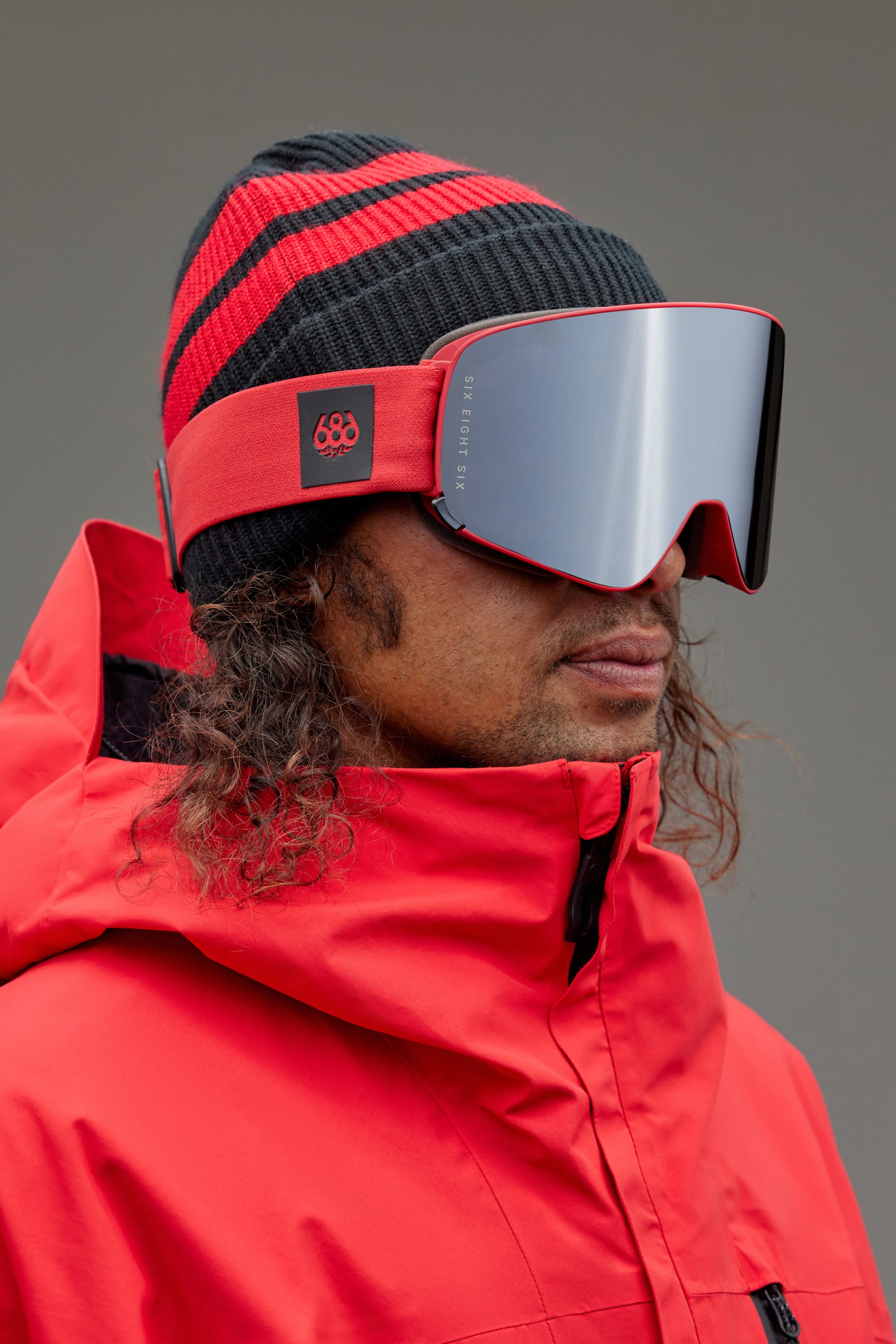 A person wearing a black and red striped beanie, 686 SMARTY Goggle by 686 with magnetic lenses, and a bright red jacket stands against a plain gray background.