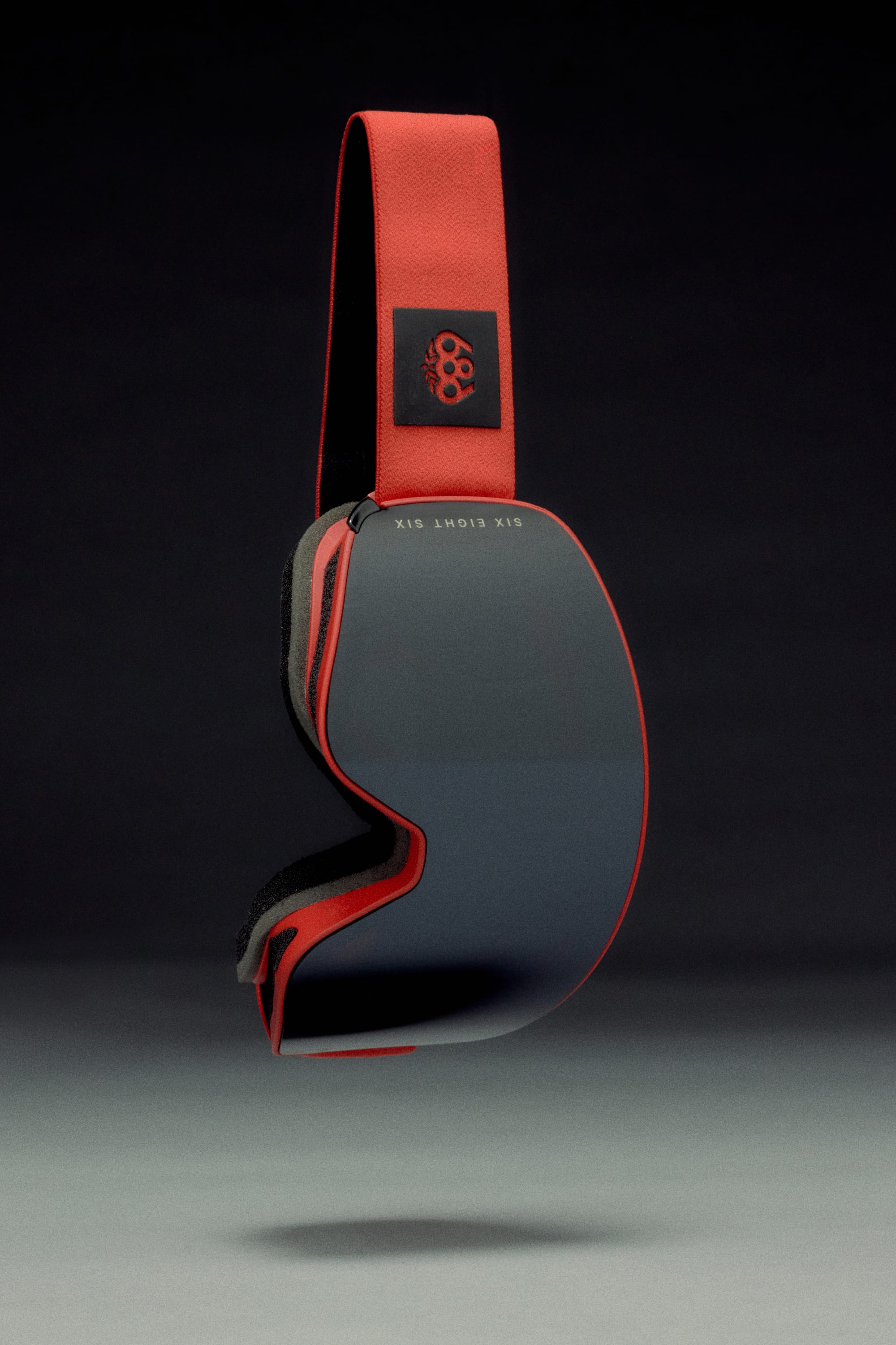 The 686 SMARTY Goggle by 686 features a modern minimalist design with a reflective dark lens, red strap, and small black emblem, suitable for all conditions and shown against a dark background.