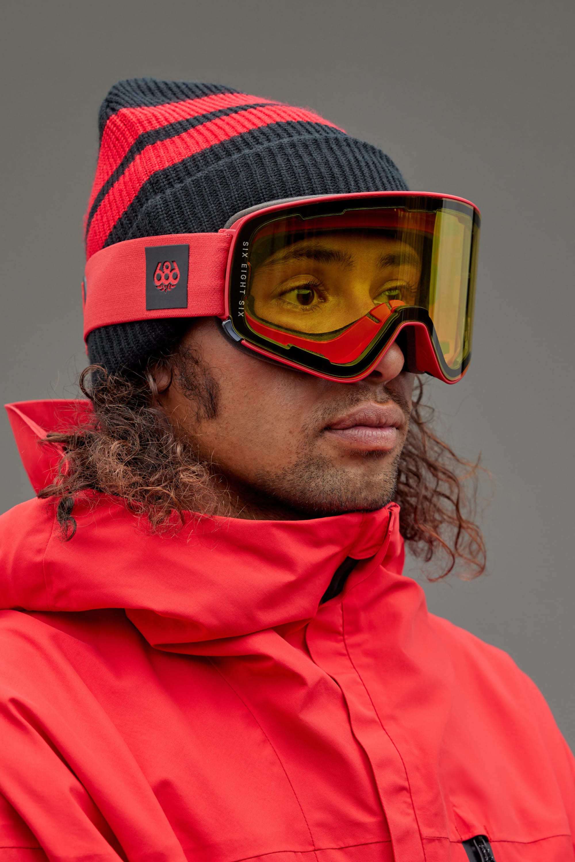 A person wearing a red and black beanie, 686 SMARTY Goggle by 686 with magnetic lenses, and a red winter jacket looks off to the side against a plain background.