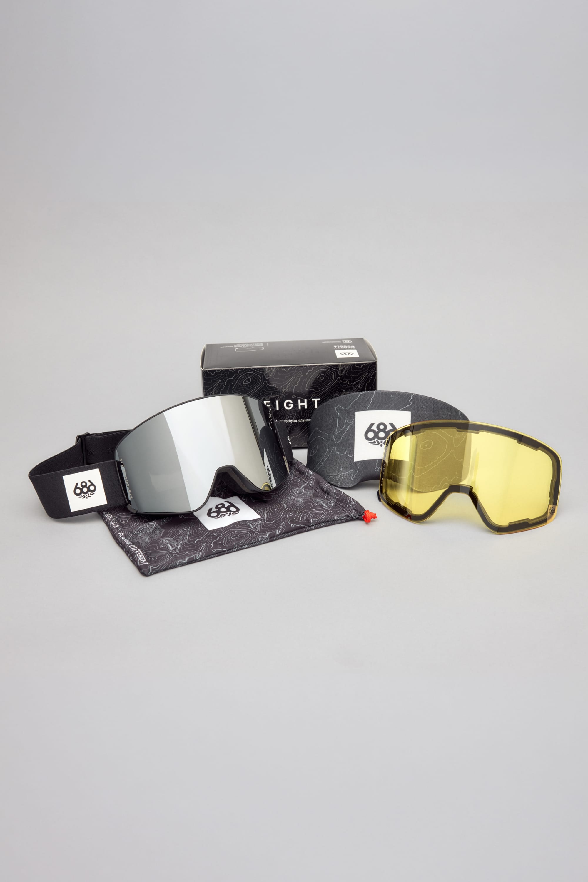 The 686 SMARTY® Magnetic Goggle by 686, featuring mirrored lenses with a black strap, comes with a yellow replacement lens, a gray pouch, and a black box—all displayed on a light gray surface.