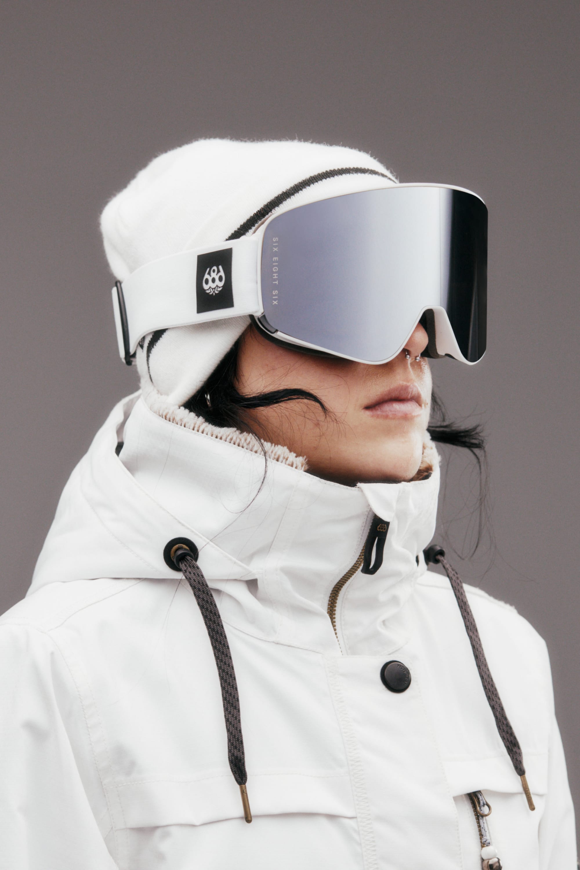 Wearing a white winter jacket, white beanie, and the 686 SMARTY Goggle by 686 with magnetic lenses, a person stands against a plain gray background—ready for all conditions.