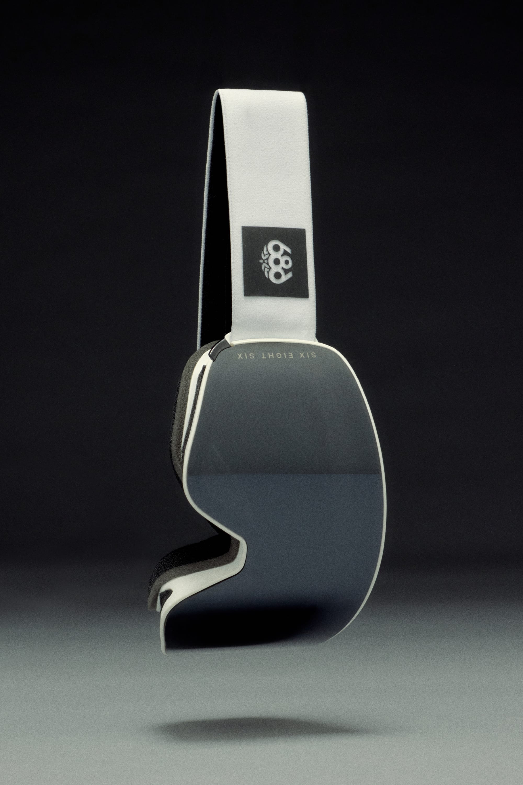 The 686 SMARTY Goggle by 686 features magnetic lenses, a wide dark lens, and a white strap with a small black logo, presented against a dark gradient background—designed for all conditions.