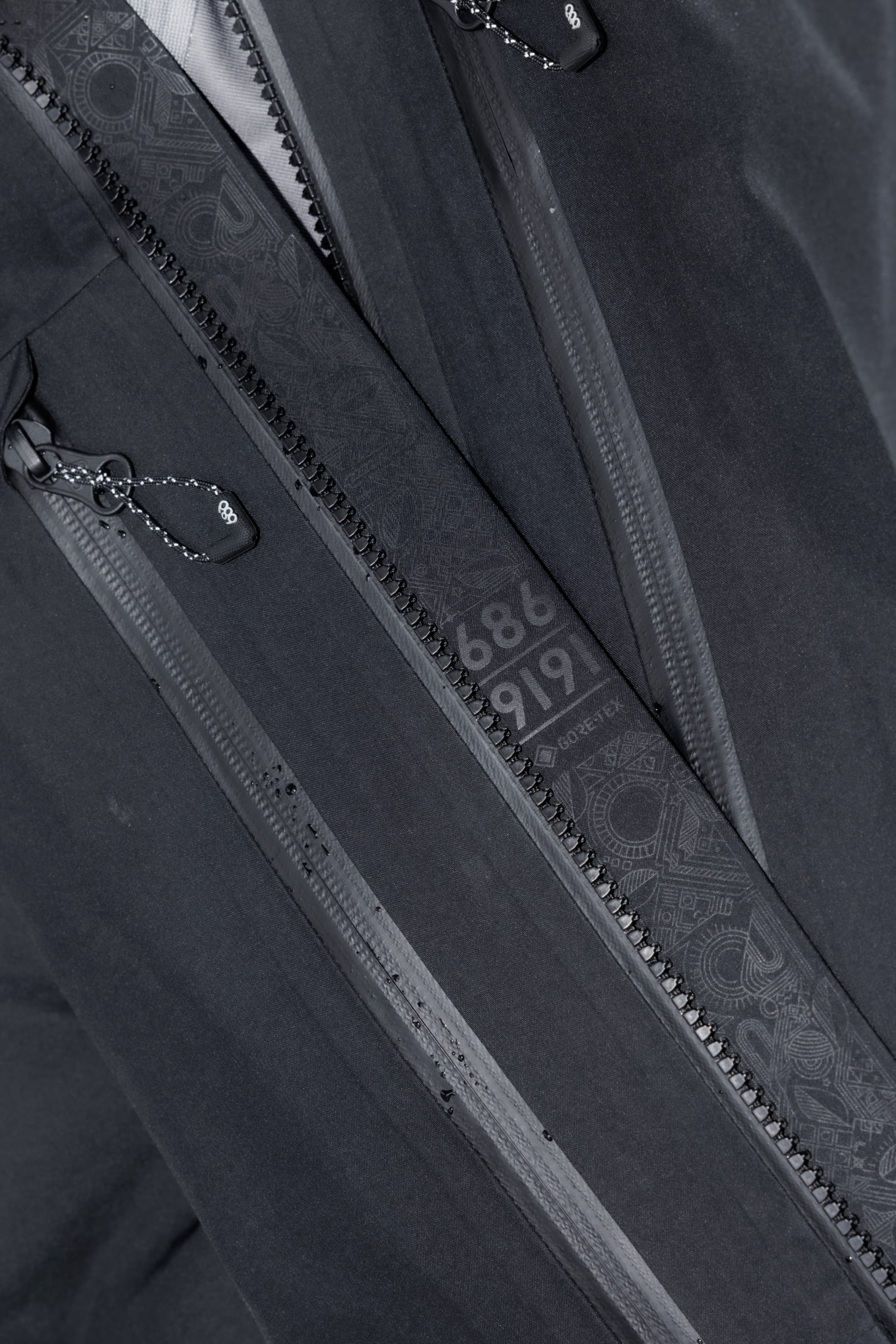 Close-up of the 686 Mens GORE-TEX 3L ATV Jacket in black, crafted from recycled fabric. Features dual zippers with decorative pull cords and an inner flap with subtle patterns plus light gray 686 and 9191 detailing.