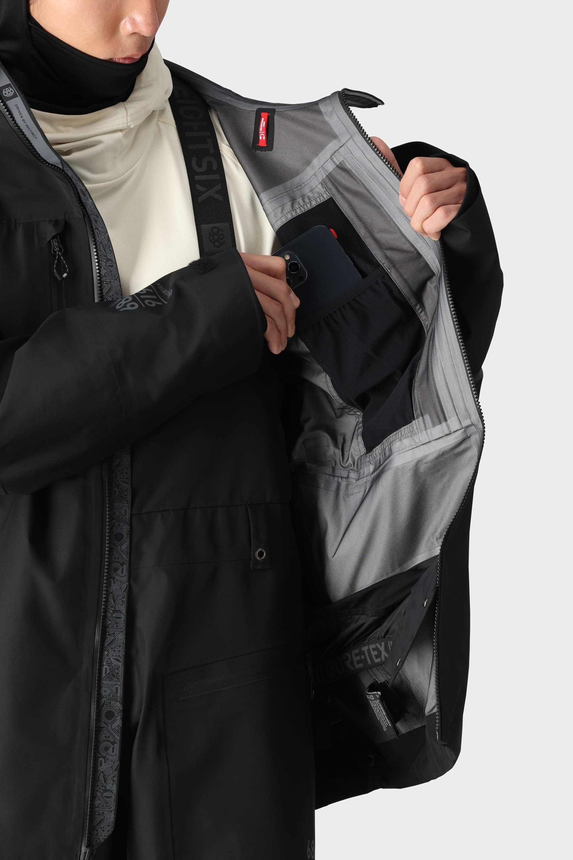 A man in the 686 Mens GORE-TEX 3L ATV Jacket opens the inner pocket, revealing organized compartments and stashes his phone. The light gray recycled lining features multiple pockets for easy organization.
