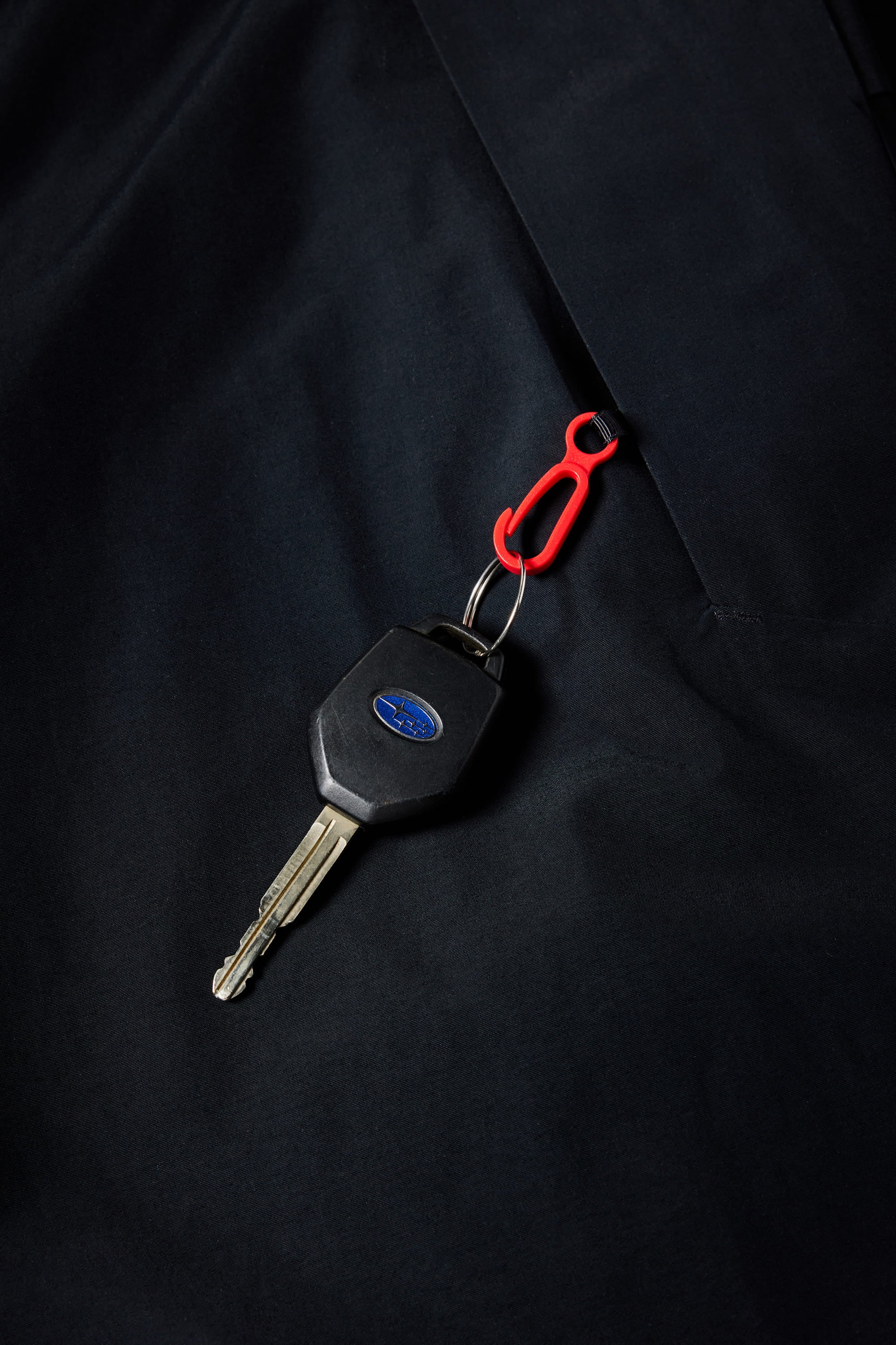 A car key with a black plastic head and blue oval logo is attached to a small red carabiner, resting on the dark, waterproof surface of a 686 Mens Waterproof Slapshot Hoody by 686.