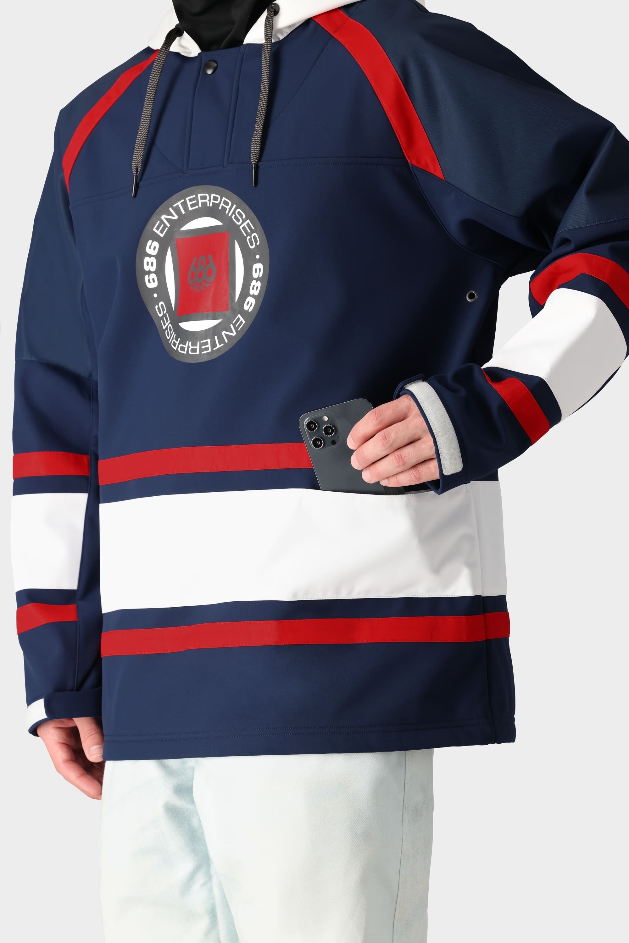 A person wears the 686 Men’s Waterproof Slapshot Hoody, featuring blue, red, and white hockey-style design with 686 ENTERPRISES on the front, holding a black smartphone in a front pocket against a plain white background.