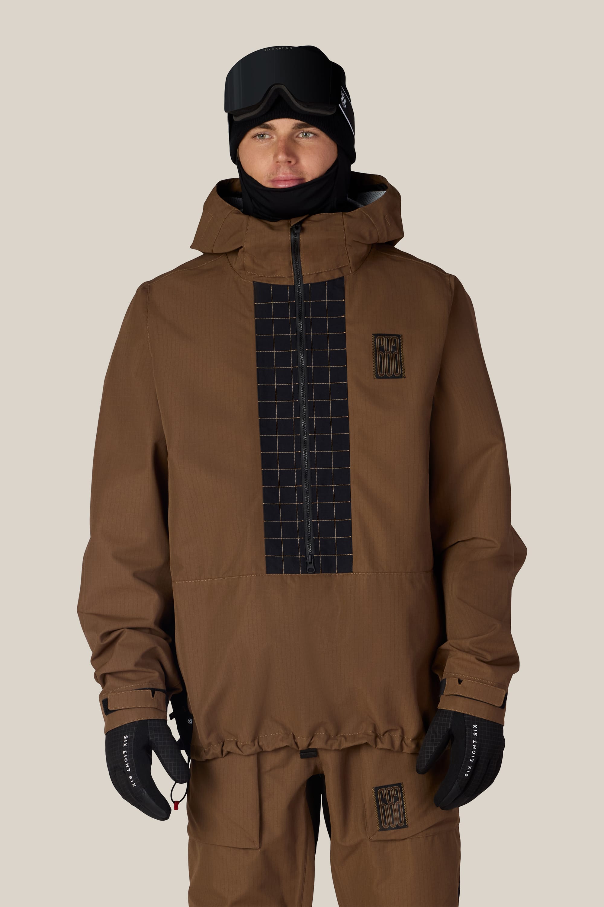A person wearing the 686 Men’s Ghost™ 2.5L Anorak by 686, featuring a brown color with black grid patterns, paired with matching pants, black gloves, and black ski goggles against a light beige background.