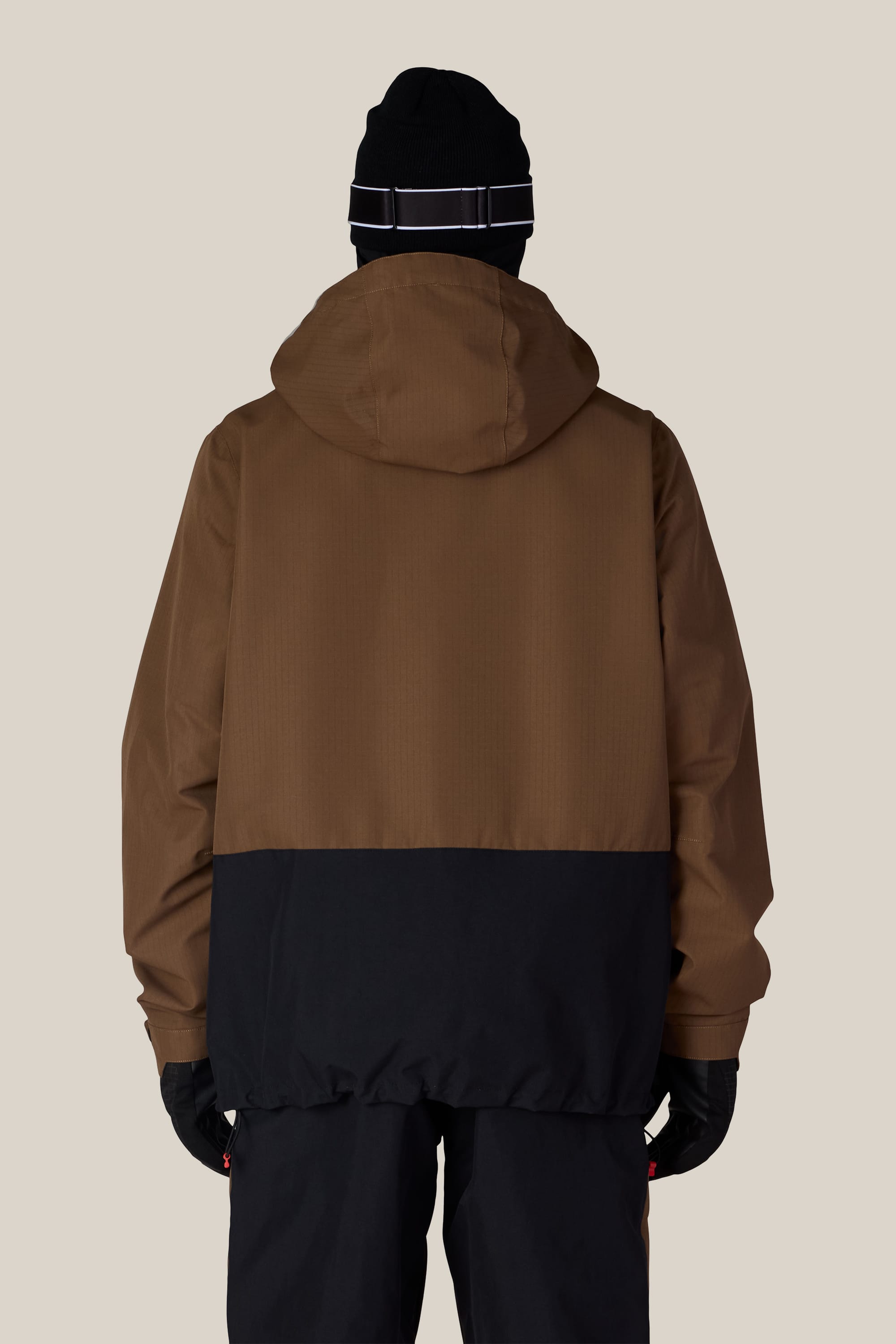 A person facing away, wearing the 686 Men’s Ghost™ 2.5L Anorak in brown and black, along with black gloves, dark pants, and a black beanie with a white stripe, stands against a plain light background.