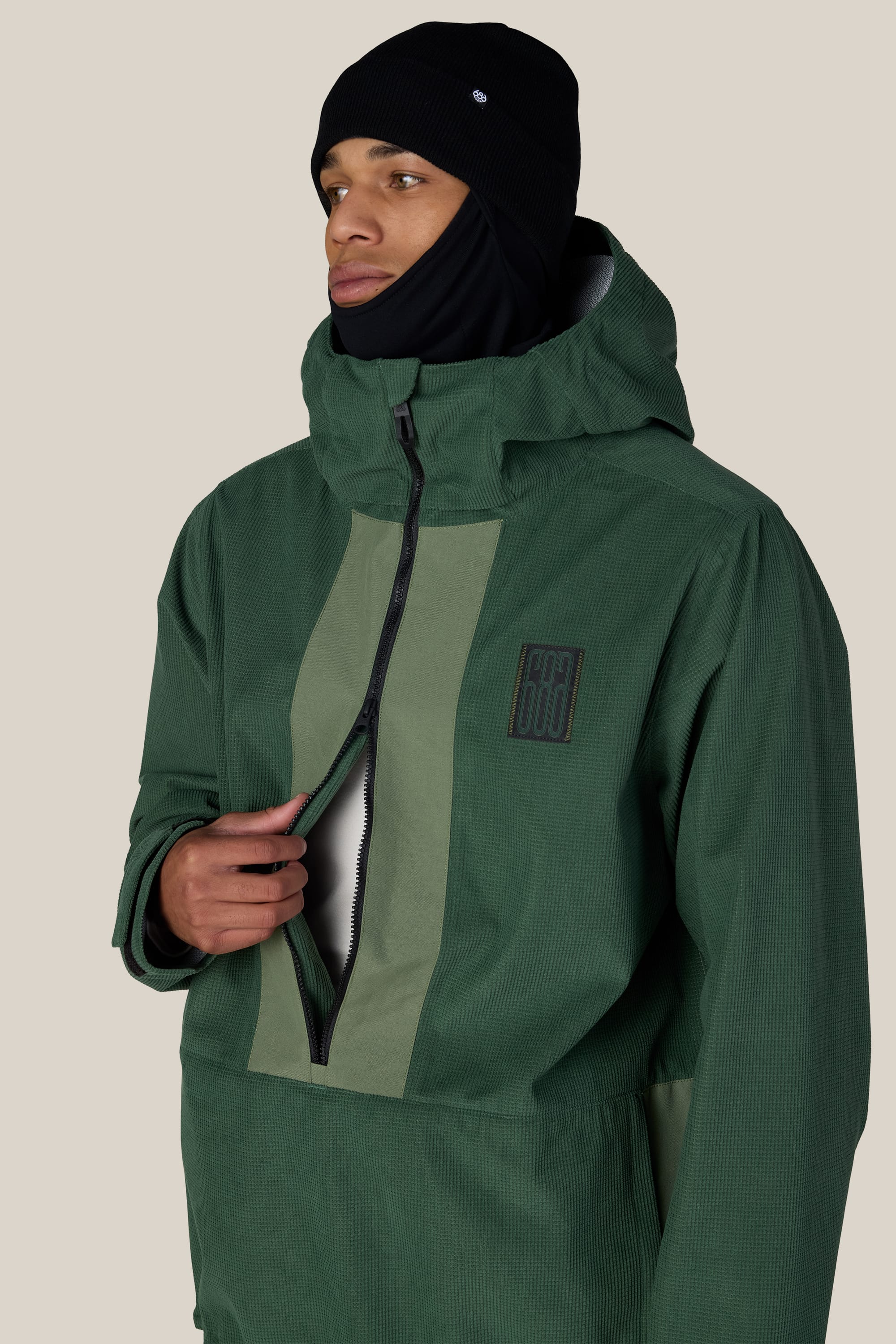 A person wearing the 686 Men’s Ghost™ 2.5L Anorak in dark green and a black balaclava looks to the side while unzipping the front of the jacket against a plain beige background.