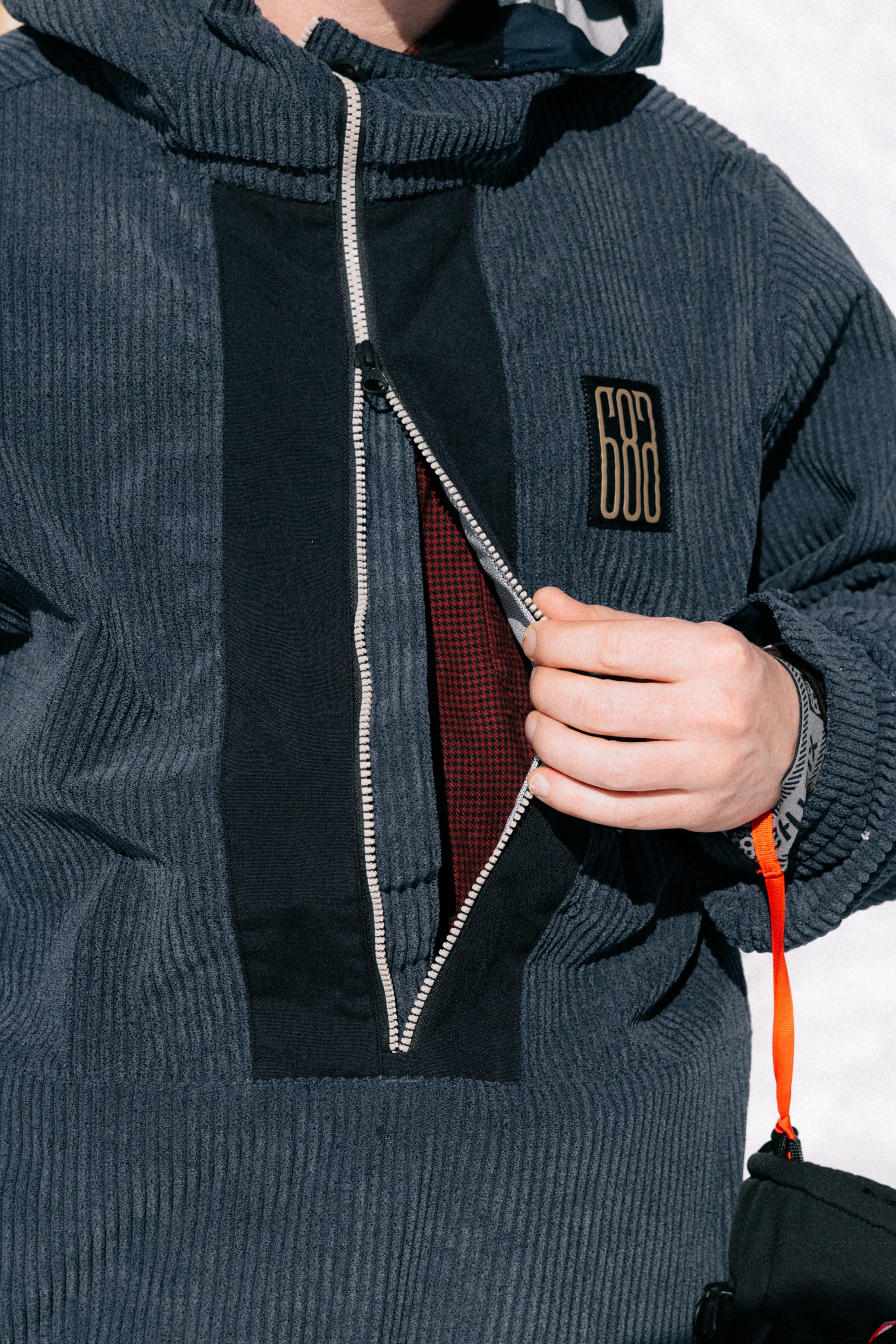 A person unzips the 686 Men’s Ghost™ 2.5L Anorak, a blue corduroy jacket featuring a black vertical stripe and a signature 686 patch, revealing its red checkered inner lining. A wristband and orange strap are visible on their wrist.