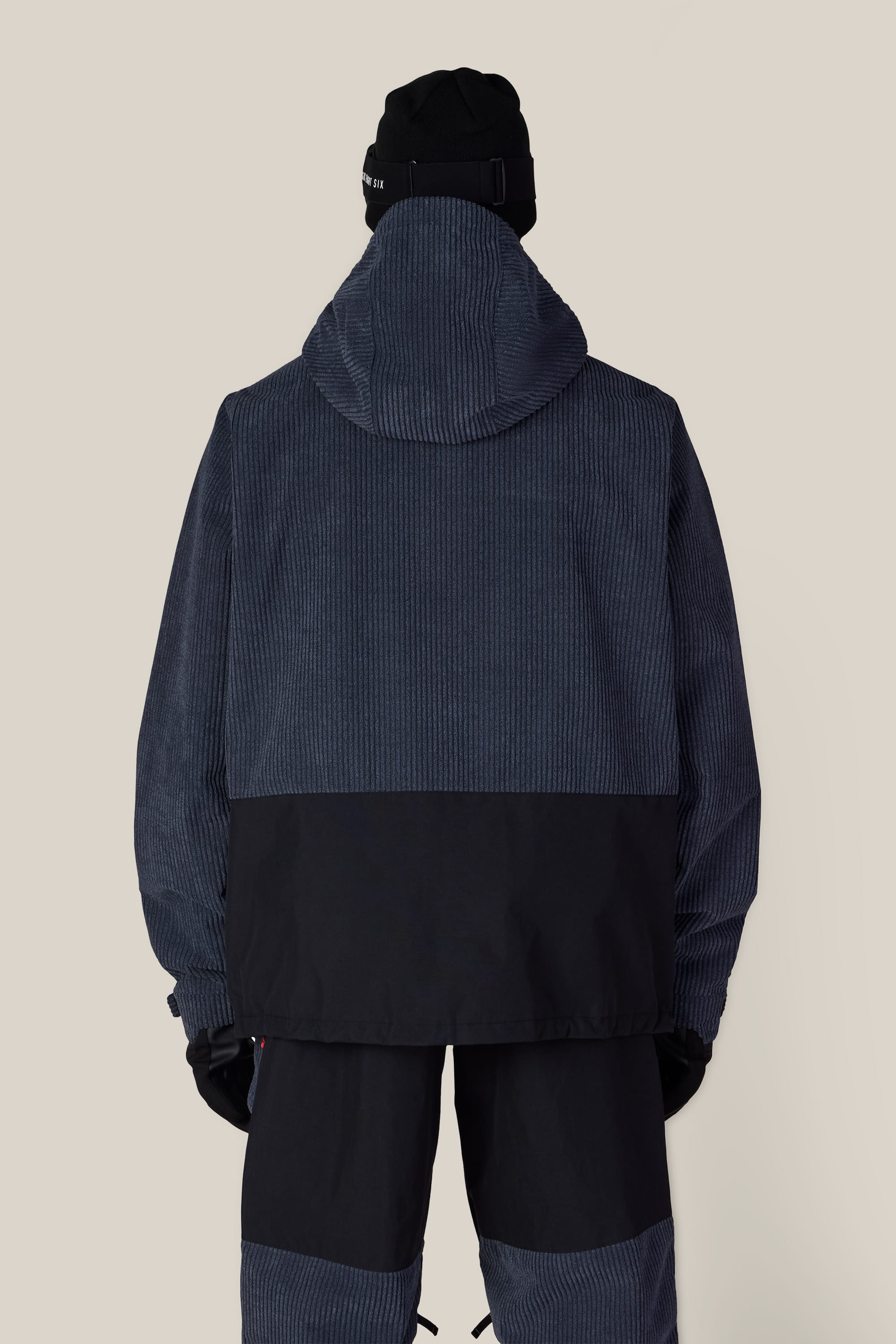 A person is seen from the back against a plain beige background, wearing the 686 Men’s Ghost™ 2.5L Anorak by 686—a dark blue corduroy hooded jacket—paired with a black beanie, black gloves, and matching pants.