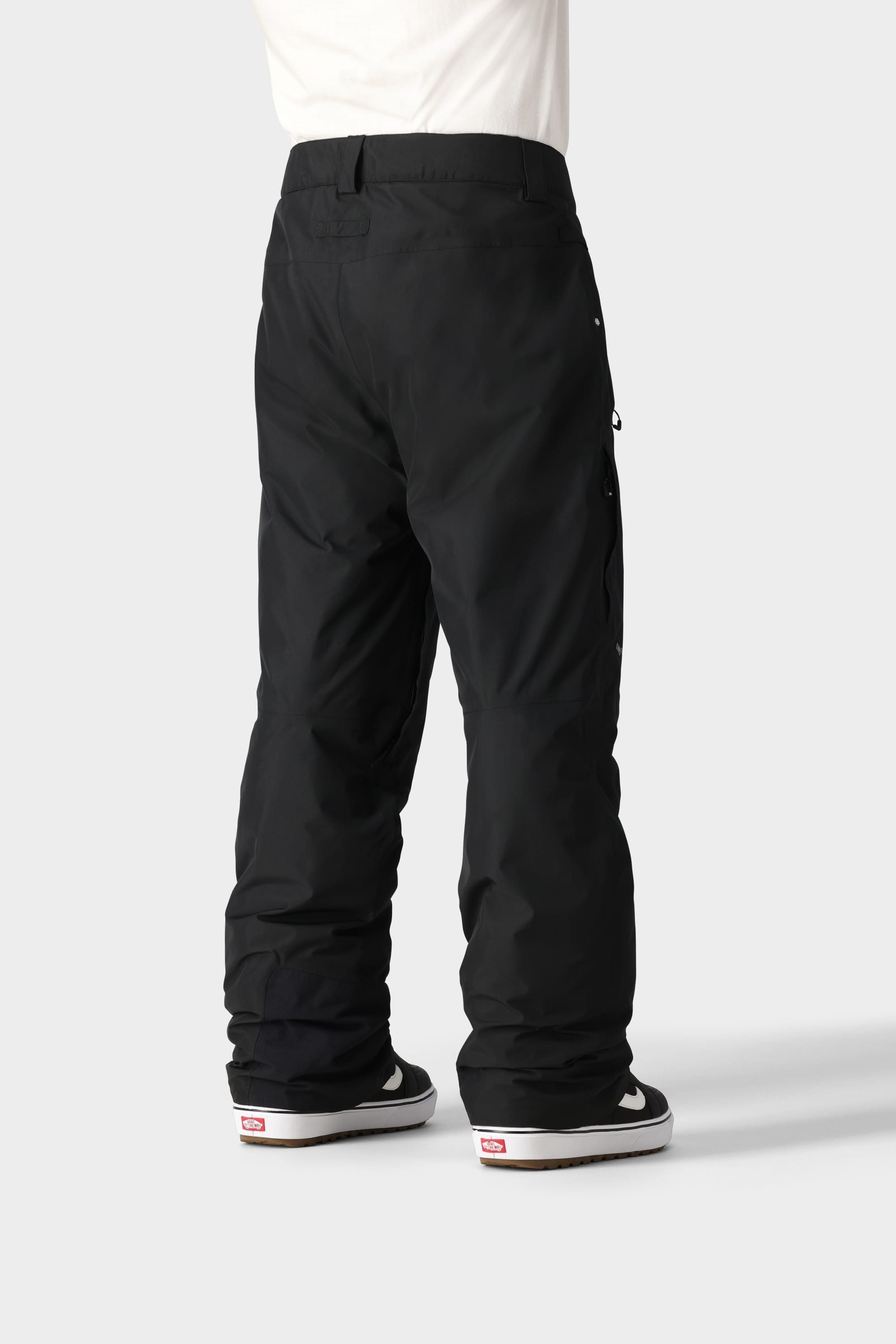 A person wearing 686 Mens GORE-TEX Core Insulated Pant and black-and-white Vans sneakers stands with their back to the camera against a light gray background, showing only the lower half of their body.