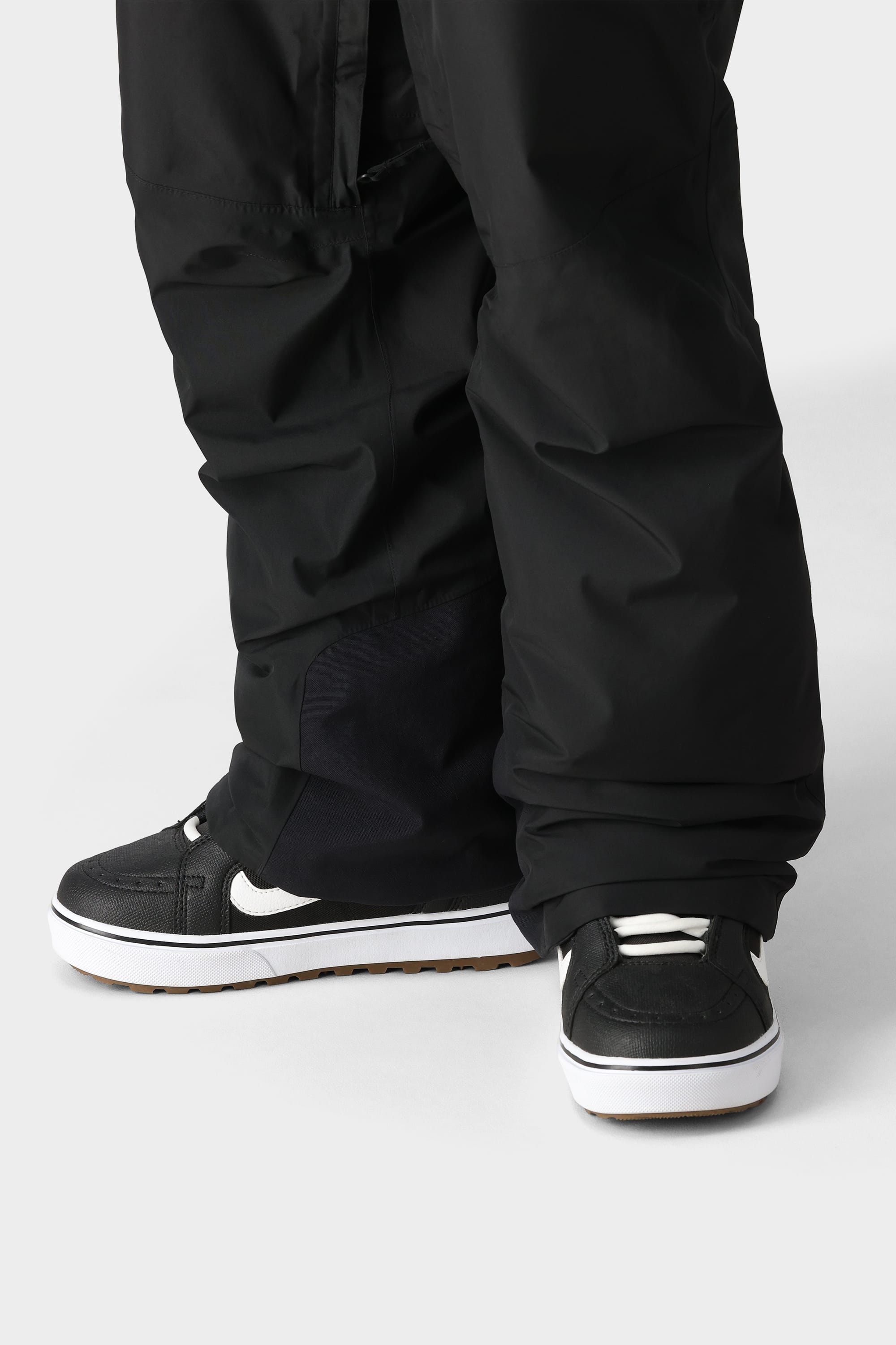 A close-up of a person wearing black baggy 686 Mens GORE-TEX Core Insulated Pants that partially cover black high-performance sneakers with white soles, standing on a light gray surface.