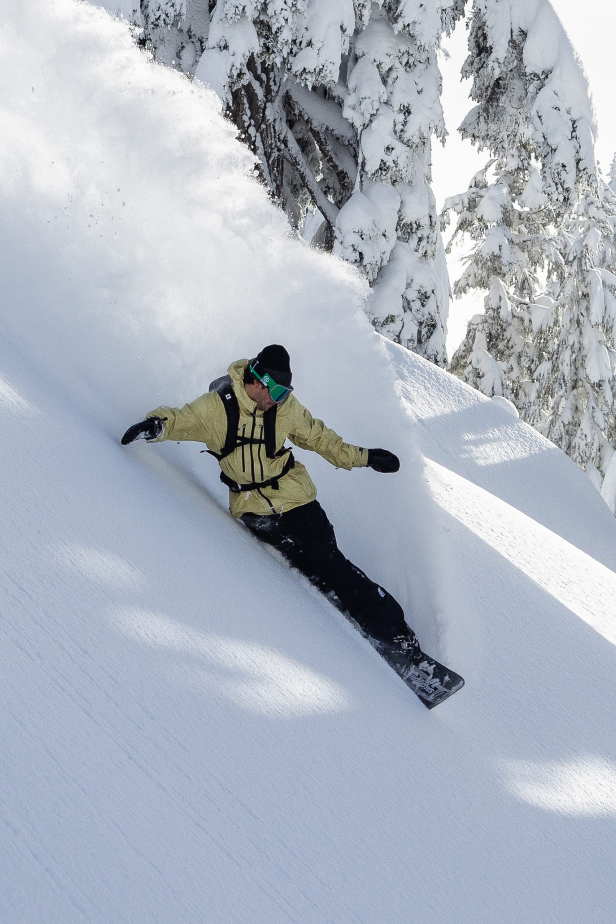 Wearing 686 Mens GORE-TEX Core Insulated Pants, a snowboarder in a yellow jacket and green goggles carves through fresh powder on a steep slope, flanked by snow-covered trees under bright daylight.