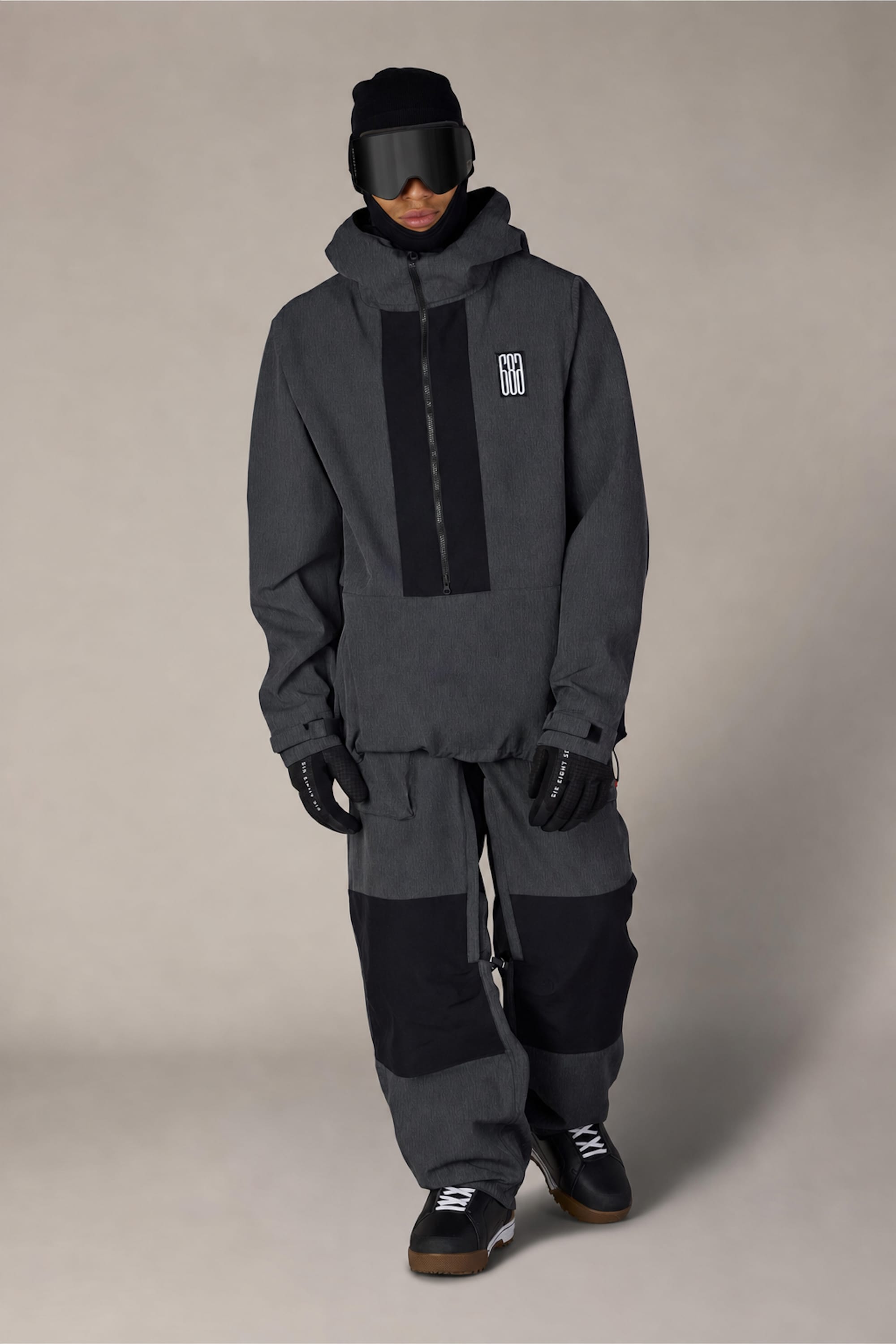 A person wearing the 686 Ghost™ 2.5L Pant from 686, paired with a dark gray and black winter outfit, gloves, a hood, beanie, and ski goggles stands against a neutral background. The jacket features a vertical zipper and displays a white logo on the chest.