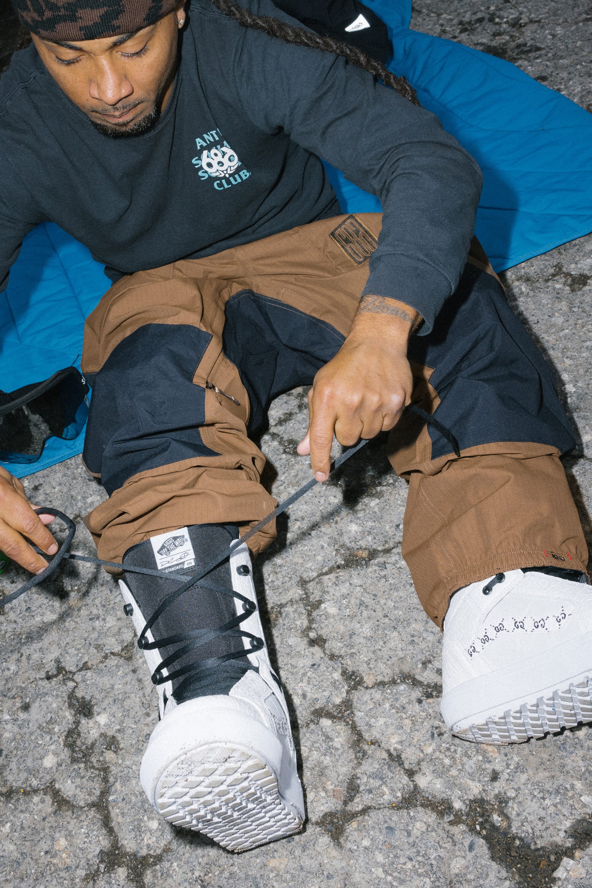 A person sits on a blue mat outdoors, tying the laces of a white boot while wearing the 686 Ghost™ 2.5L Pant by 686 and a black long-sleeve shirt; the ground beneath them is cracked pavement.