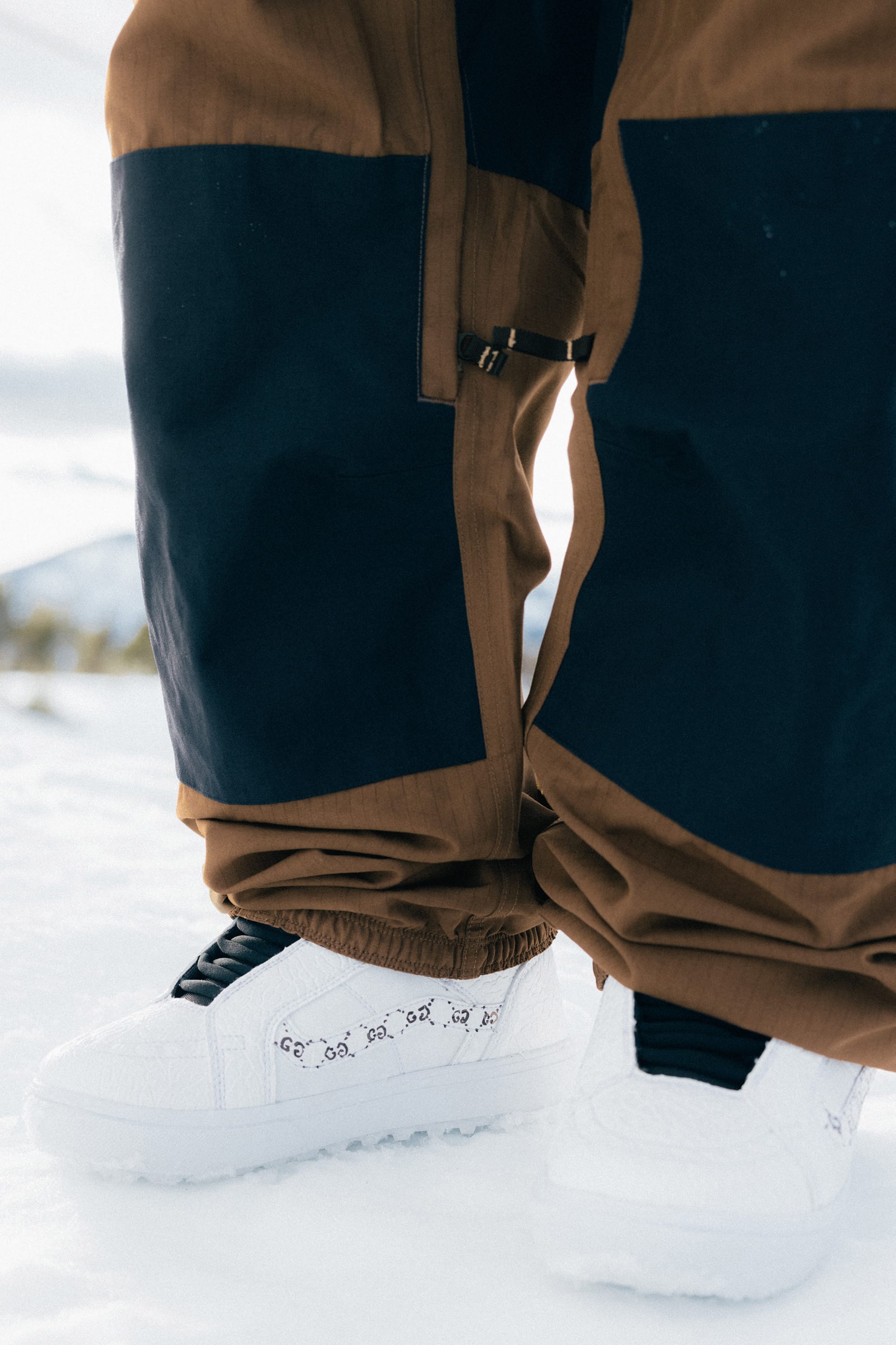 A person wearing the 686 Ghost™ 2.5L Pant by 686 stands in fresh snow, paired with white sneakers featuring a unique black side pattern. The wintry scene appears cold and crisp.