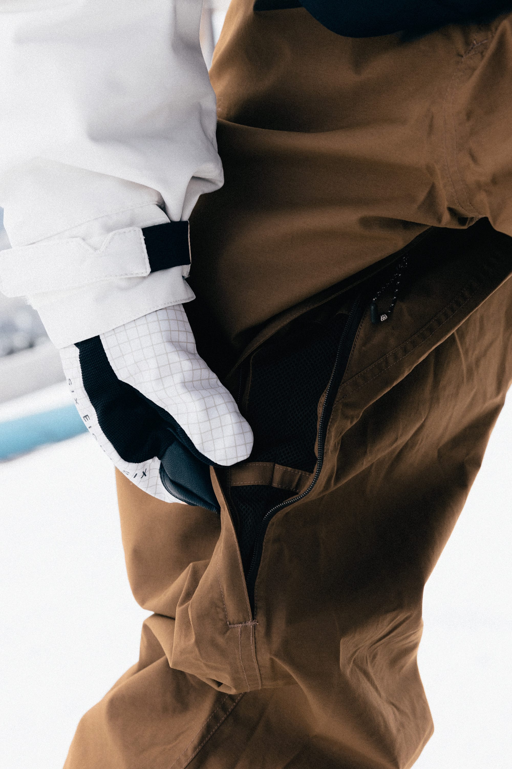 Wearing the 686 Ghost™ 2.5L Pant, a person in white gloves and a white jacket opens a zippered pocket while standing on snow.