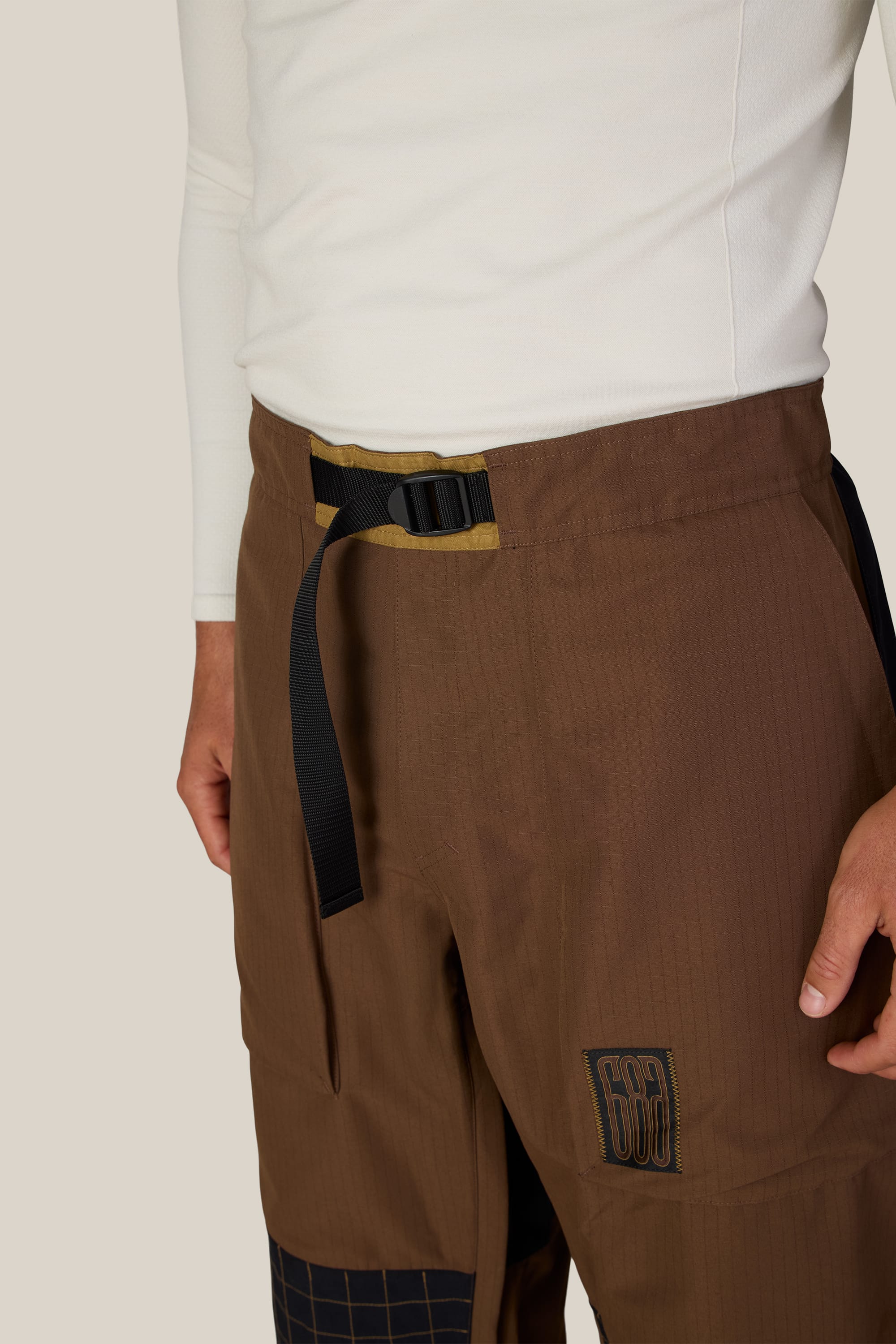 A person is wearing the 686 Ghost™ 2.5L Pant by 686, featuring a stitched patch and grid pattern accents, styled with a black belt and a white long-sleeve shirt. Only the lower torso and one hand are visible.