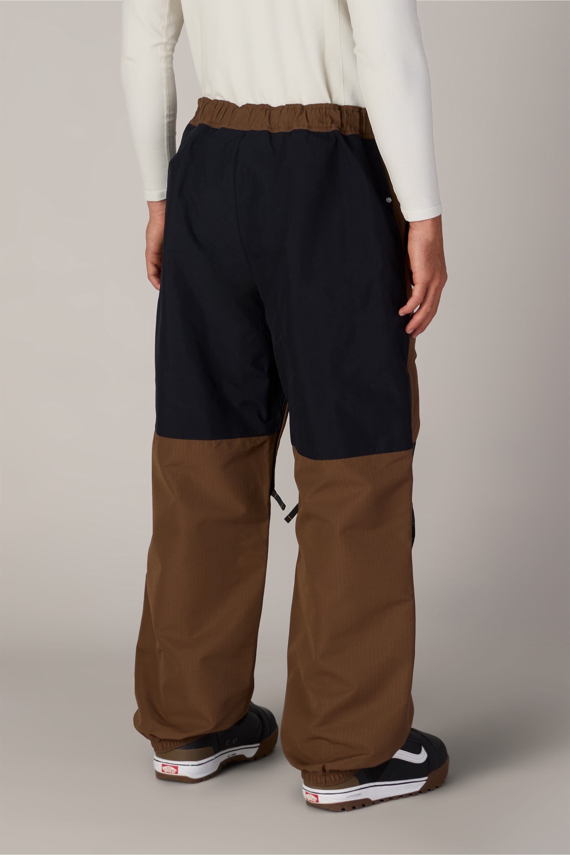 A person is seen from behind, standing on a light gray floor and wearing the 686 Ghost™ 2.5L Pant by 686, which features black upper and brown lower sections, paired with a white long-sleeve shirt and black sneakers.