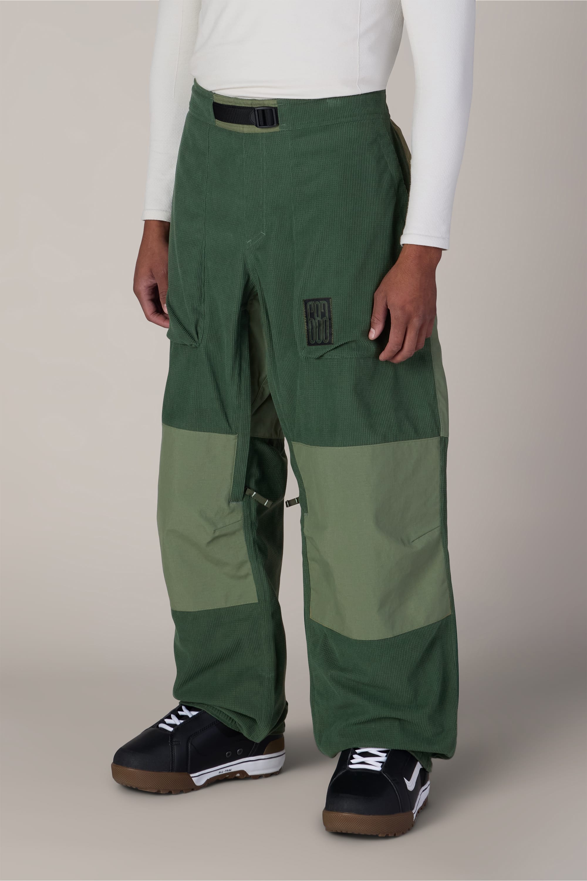 A person is wearing the 686 Ghost™ 2.5L Pant by 686—two-tone green pants with large front pockets—paired with a black belt, black sneakers with white soles, and a long-sleeve white shirt. The background is plain light gray.