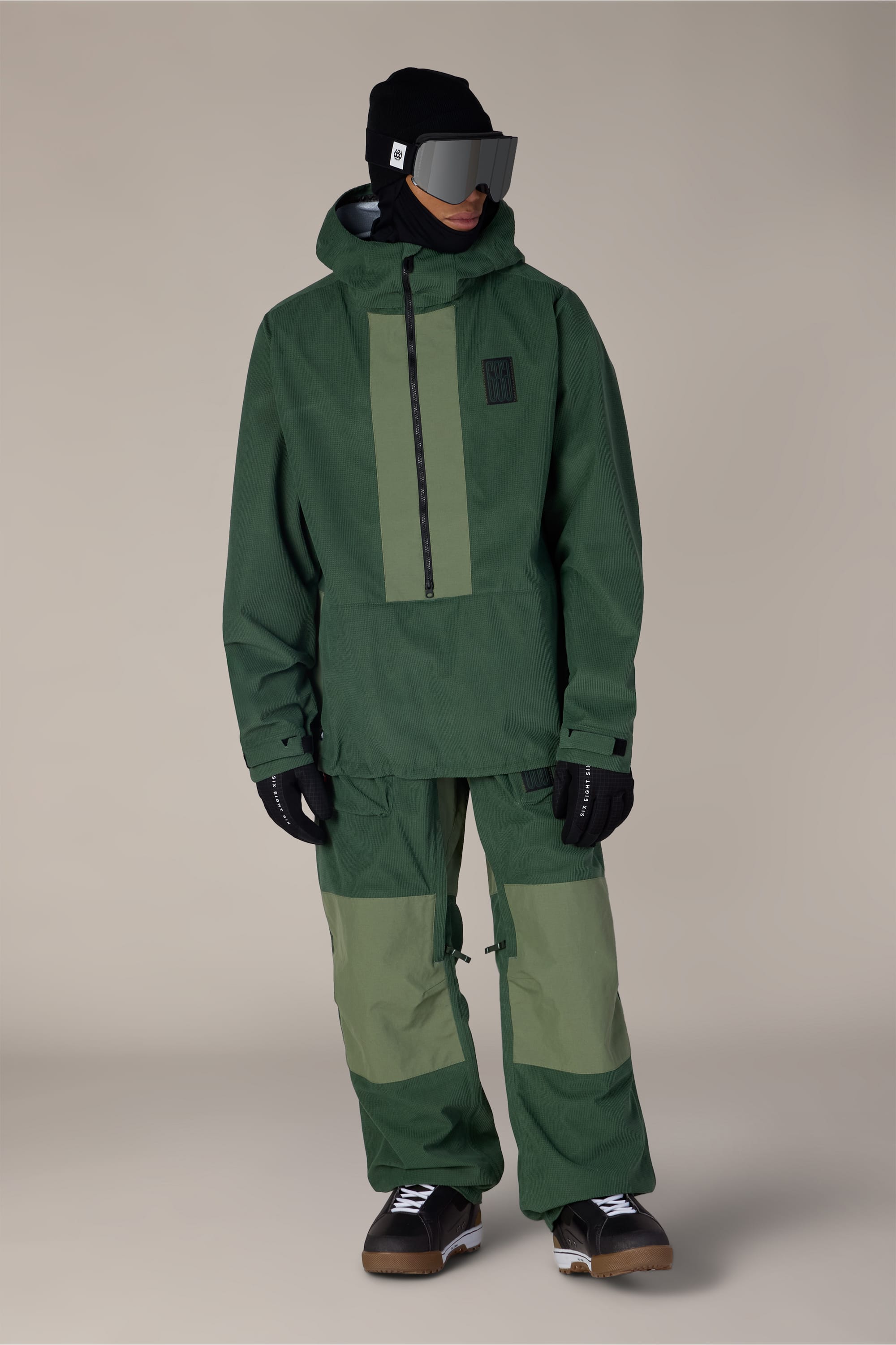 A person stands against a plain beige background wearing a green hooded ski jacket, the 686 Ghost™ 2.5L Pant in matching green by 686, black gloves, black boots, a black beanie, and large ski goggles.