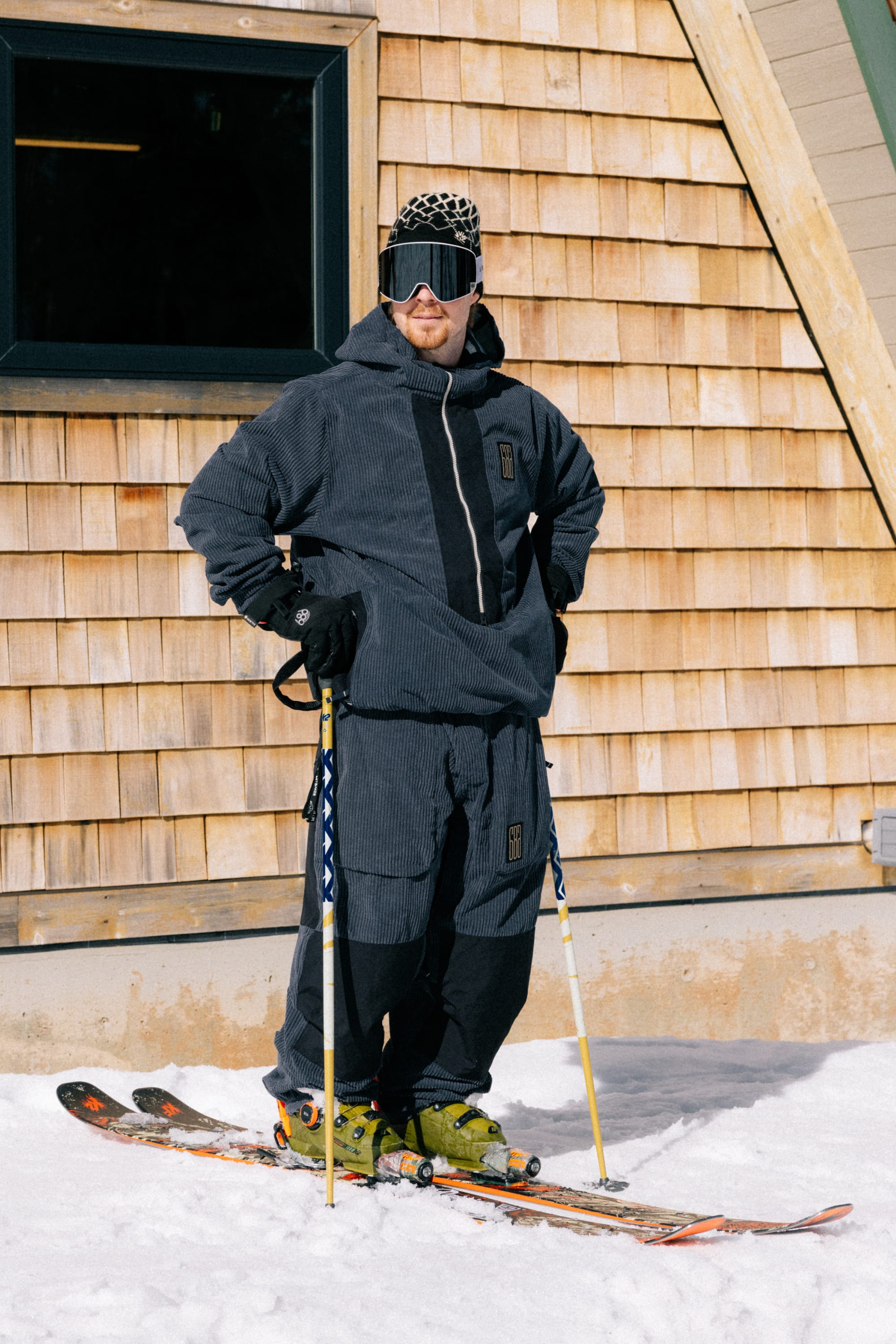 A skier wearing the 686 Ghost™ 2.5L Pant by 686, along with a dark ski suit, green ski boots, black gloves, goggles, and a patterned hat stands on snow with poles in front of a wooden building.