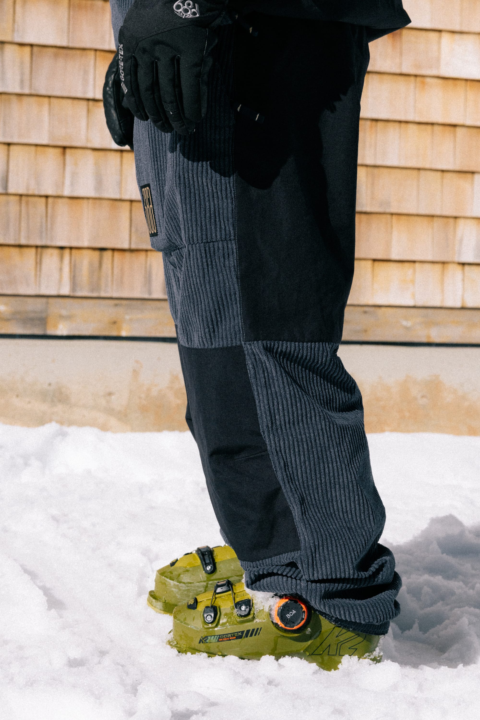 A person standing in snow wears the 686 Ghost™ 2.5L Pant, black gloves, and bright green ski boots, with a wooden wall in the background.