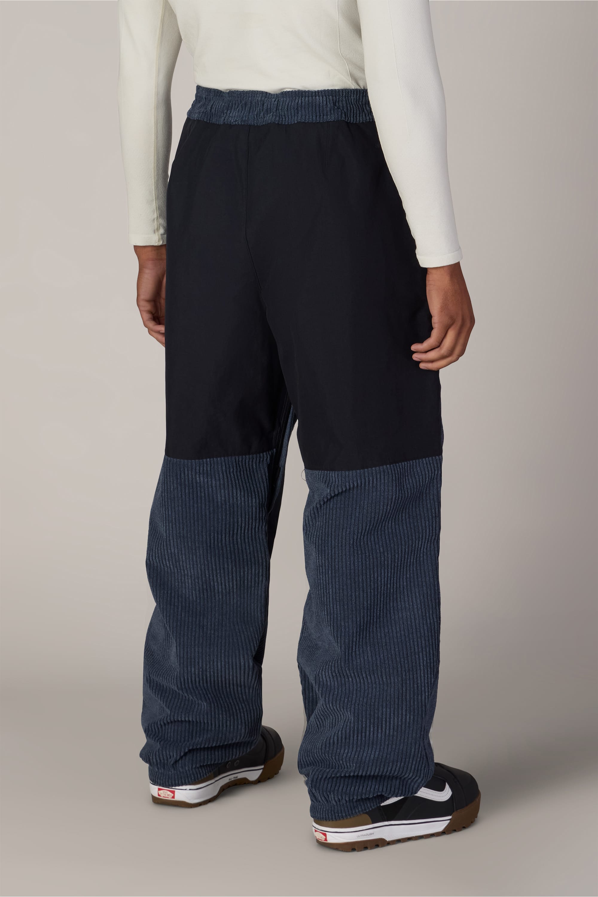 A person is standing facing away from the camera against a neutral background, wearing a white long-sleeve shirt, black sneakers with white accents, and the 686 Ghost™ 2.5L Pant by 686 in black and blue corduroy.