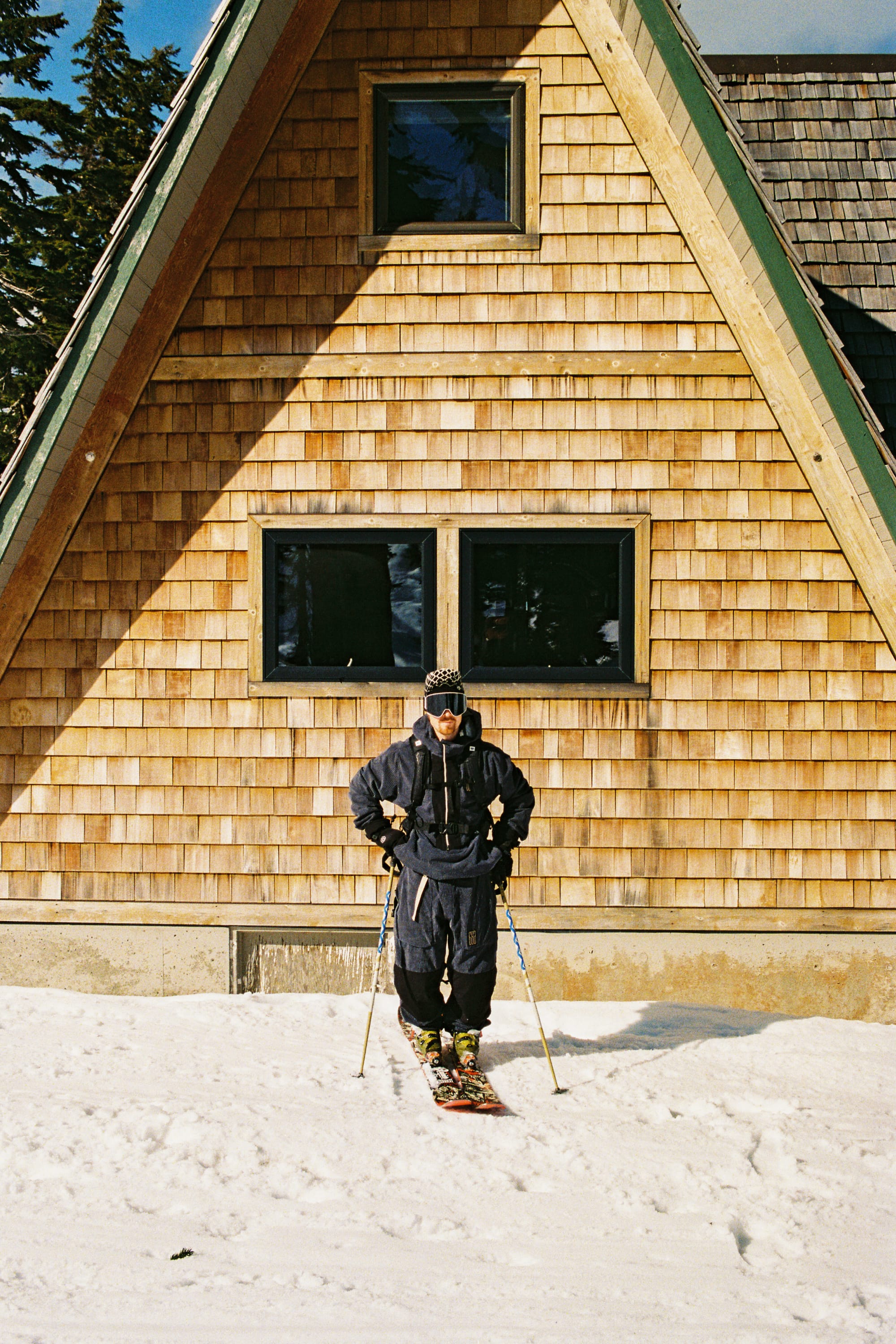 Wearing the 686 Ghost™ 2.5L Pant by 686, a person in ski gear stands on snow in front of a wooden A-frame cabin, holding ski poles, with trees visible to the left.